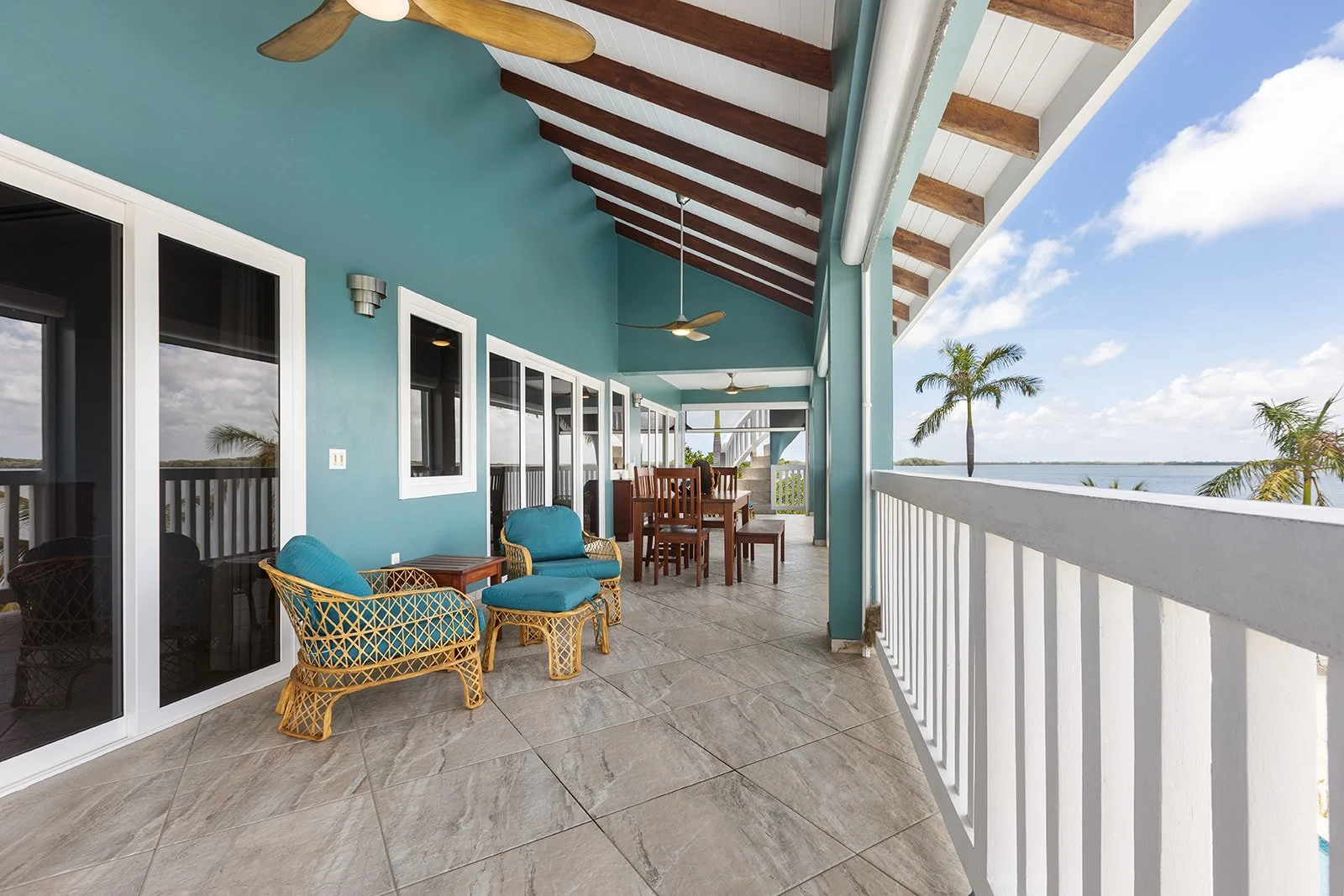 A spacious balcony with teal walls and white trim, furnished with wicker chairs with teal cushions and a dining table, overlooking palm trees and water under a partly cloudy sky.