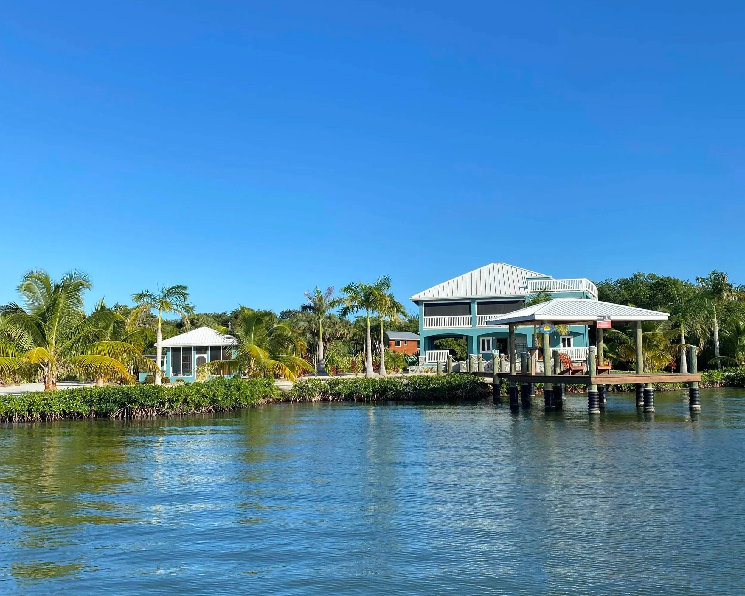 Waterfront scene with a house on stilts, surrounded by palm trees and a dock extending into the water under a clear blue sky.