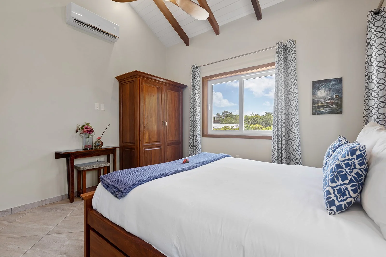 A tidy bedroom with a large bed having white linens and blue patterned pillows, a wooden wardrobe, a small desk with a flower arrangement, a window with patterned curtains showing a view of trees and sky, and a picture hanging on the wall.