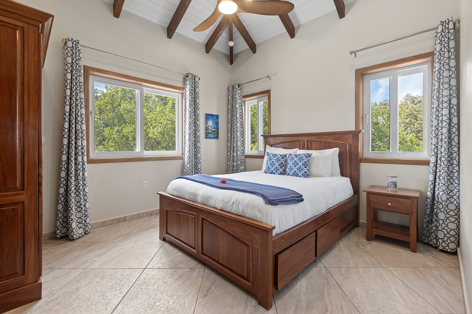Bedroom with wooden bed frame, white bedding, blue decorative pillows, nightstand with glass jar, patterned curtains, large windows showing green trees, and a ceiling fan with wooden blades.