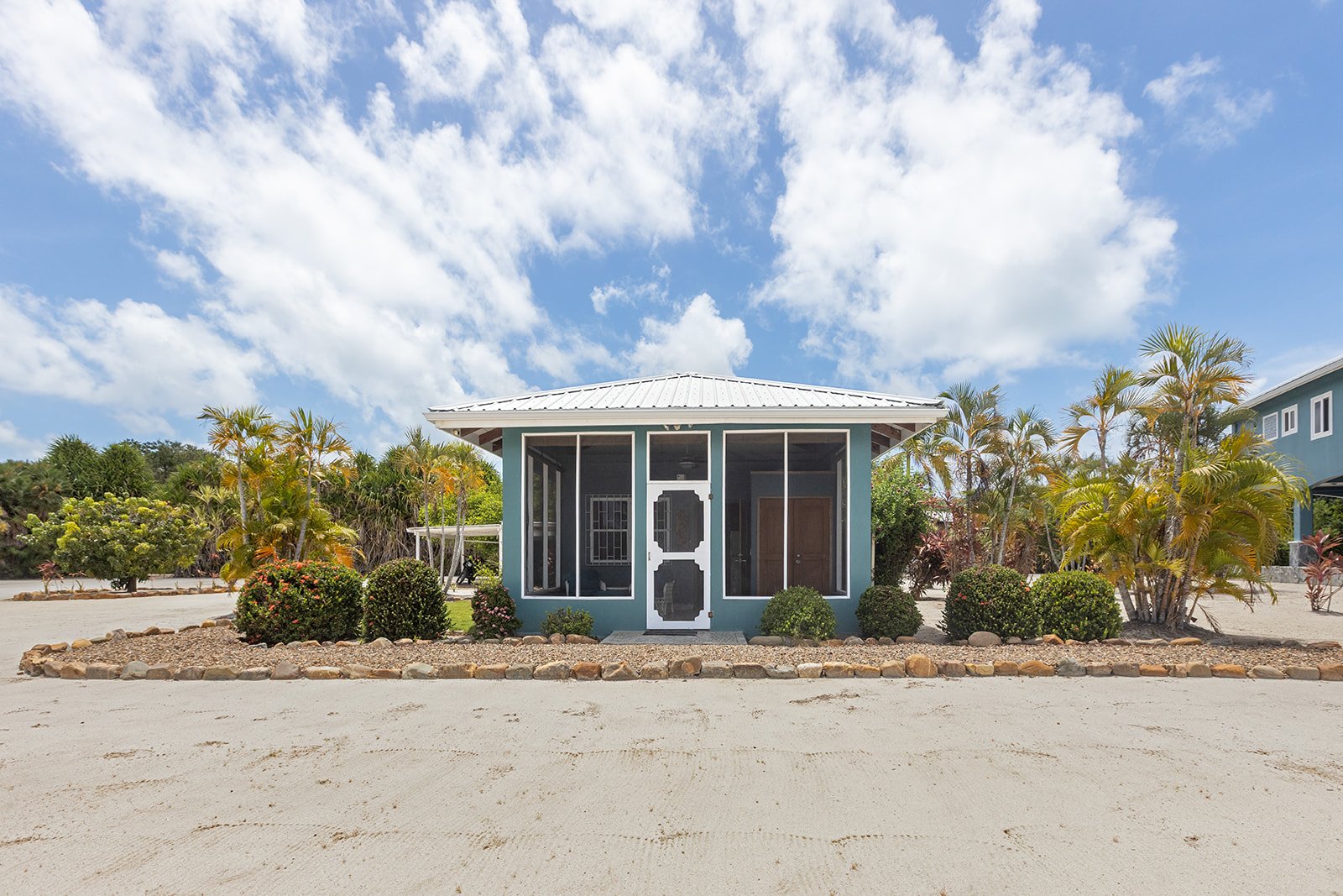 Small house with screened porch, light blue exterior, white roof, sand driveway, surrounded by tropical plants and trees, under a partly cloudy sky.