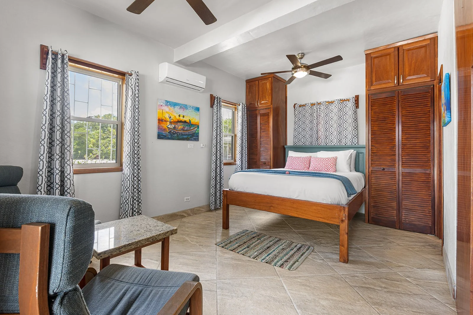 A bedroom with a wooden bed, two windows with patterned curtains, two wooden closets, a ceiling fan, a colorful painting of a boat at sunset, a small rug, and a sitting area with chairs and a table.