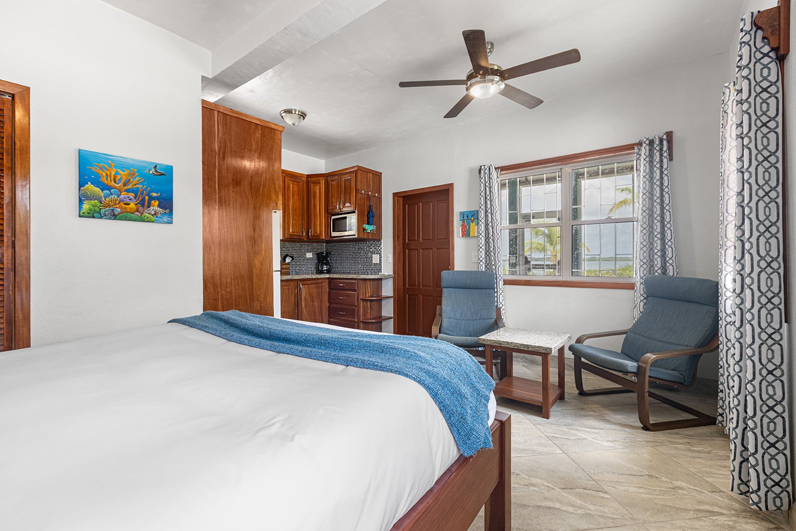 Hotel room with a bed, two blue chairs, a small table, a kitchenette with wooden cabinets, and a large window with patterned curtains.