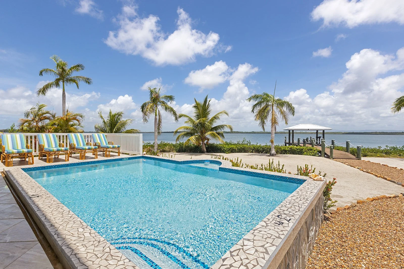 A swimming pool with striped lounge chairs on a deck, palm trees, and a view of a lake under a cloudy blue sky.