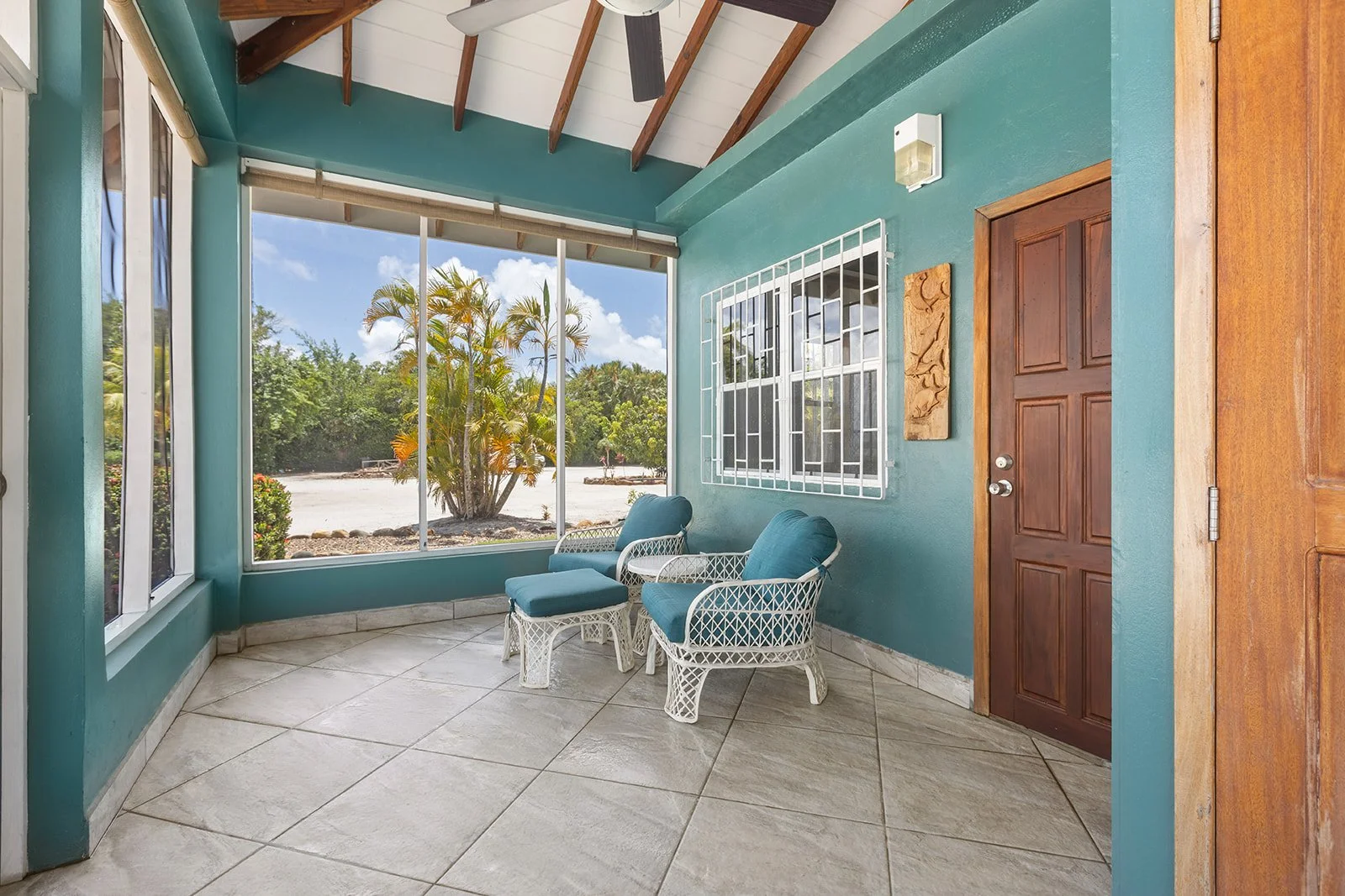 Enclosed porch with teal walls, two white wicker chairs with blue cushions, a small white table, large exterior window, wooden door, and tropical greenery outside.