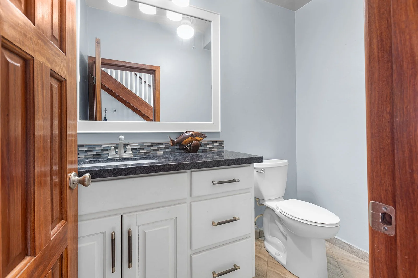 A small bathroom with a white vanity, granite countertop, a rectangular mirror, and a toilet. Wooden door and frame visible, with a staircase seen in the mirror reflection.