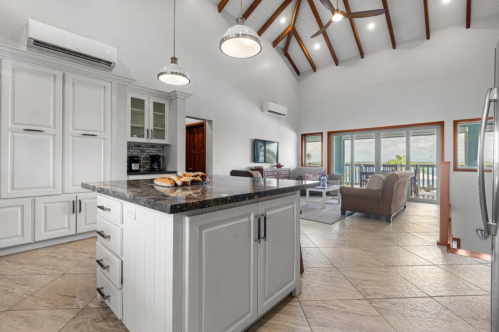 Open-concept kitchen and living room with high vaulted ceilings, white cabinetry, granite island, and large sliding glass doors leading to a balcony with ocean view.