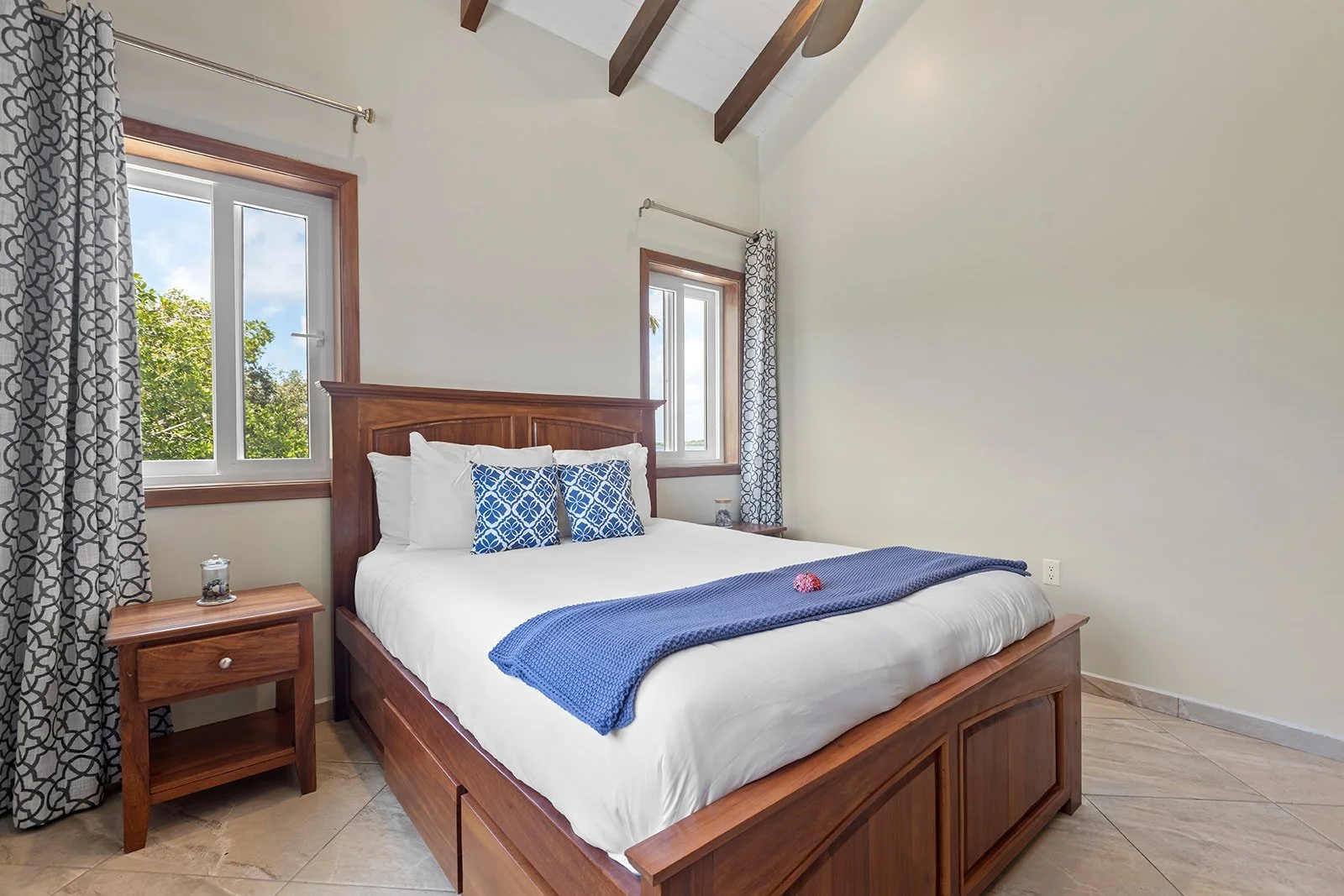 A neatly made bed with white linens, decorated with two blue patterned pillows and a blue throw blanket, in a bedroom with cream walls, wood furniture, and two windows with patterned curtains.