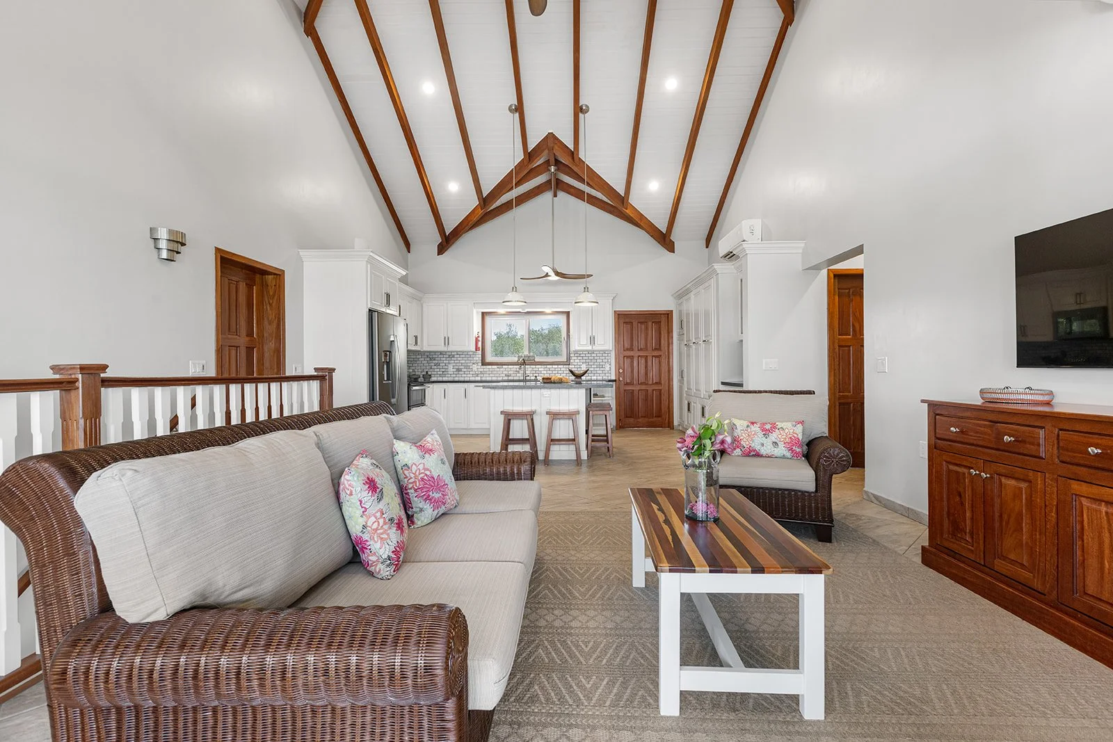 Open-concept living room and kitchen with vaulted ceiling, wooden beams, white walls, beige sofa with floral pillows, wooden coffee table with a flower vase, kitchen with white cabinets, gray backsplash, stainless steel appliances, and a window overl