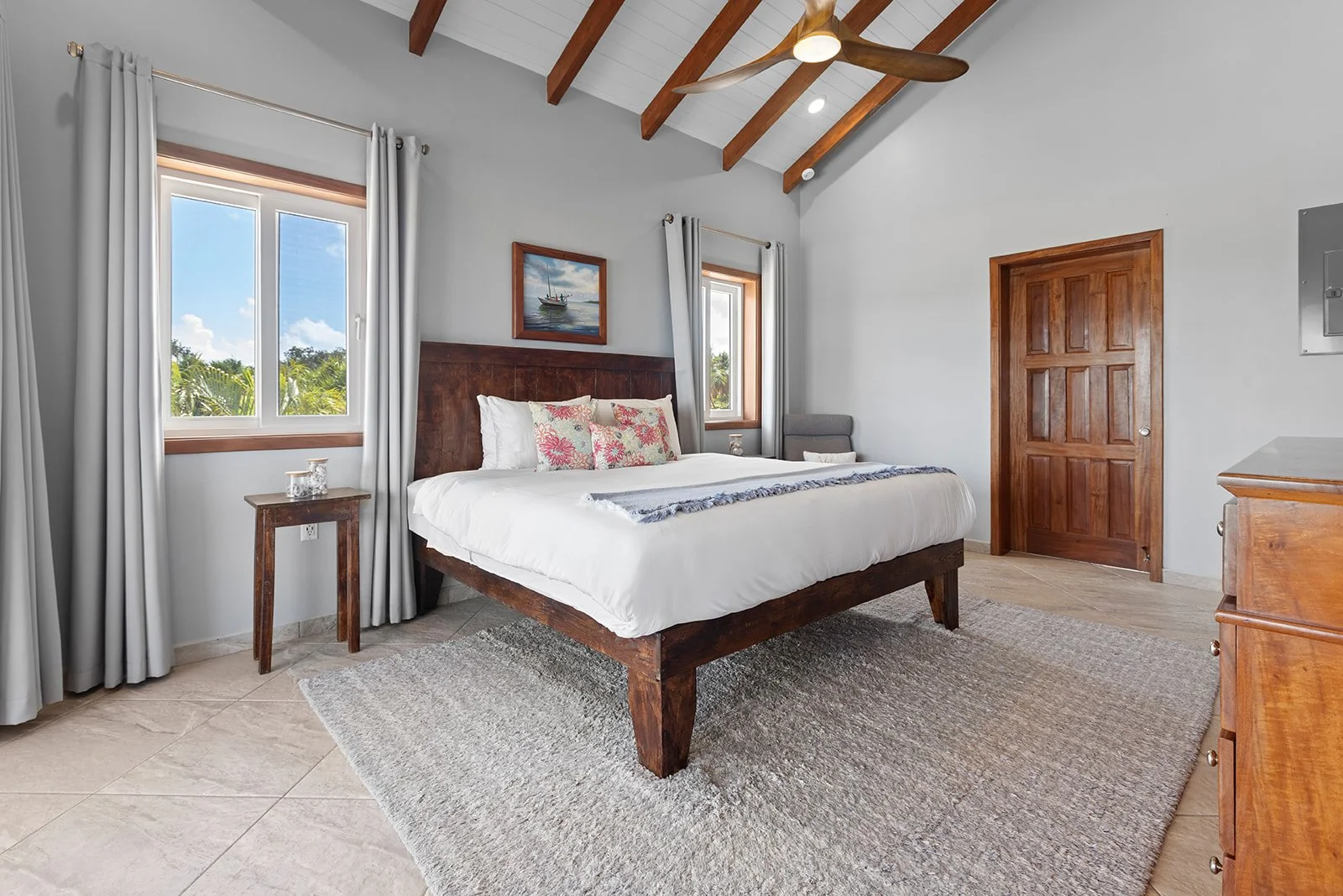 A bedroom with a large wooden bed, white bedding, and decorative pillows, flanked by two windows with light gray curtains, wooden furniture, a ceiling fan, and a beige rug on the tiled floor.