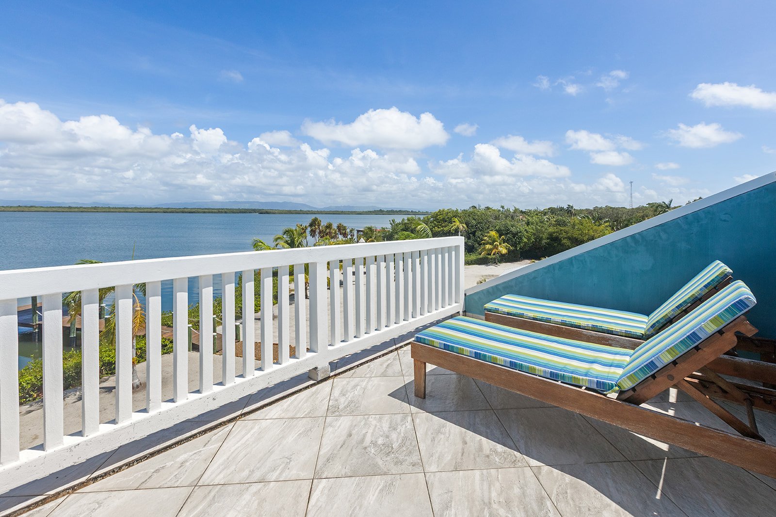 Two lounge chairs with striped cushions on a balcony overlooking a large body of water and a distant shoreline with trees and clouds in the sky.
