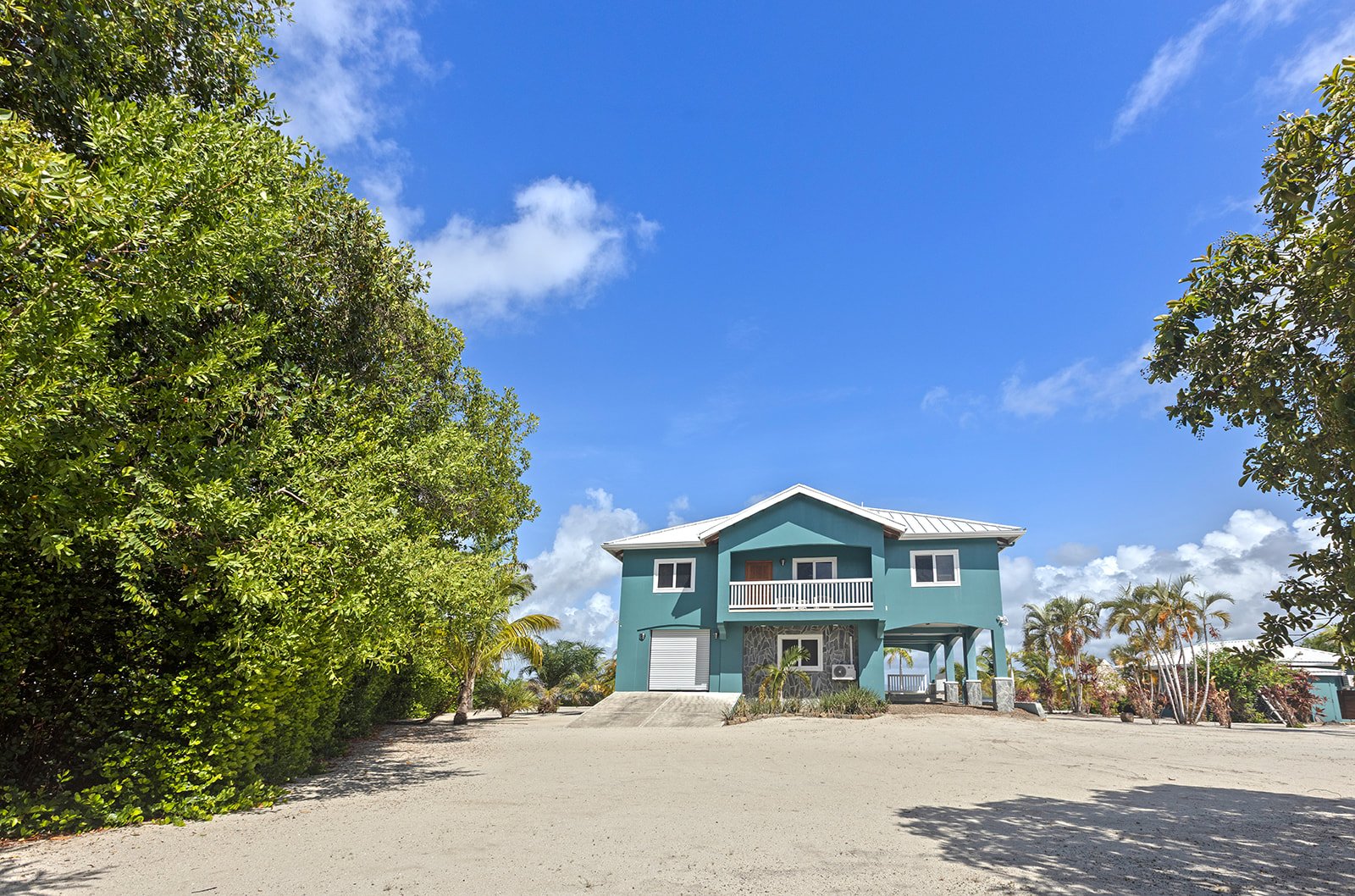 A two-story blue house with a white roof and front balcony, surrounded by greenery and palm trees, under a partly cloudy blue sky.