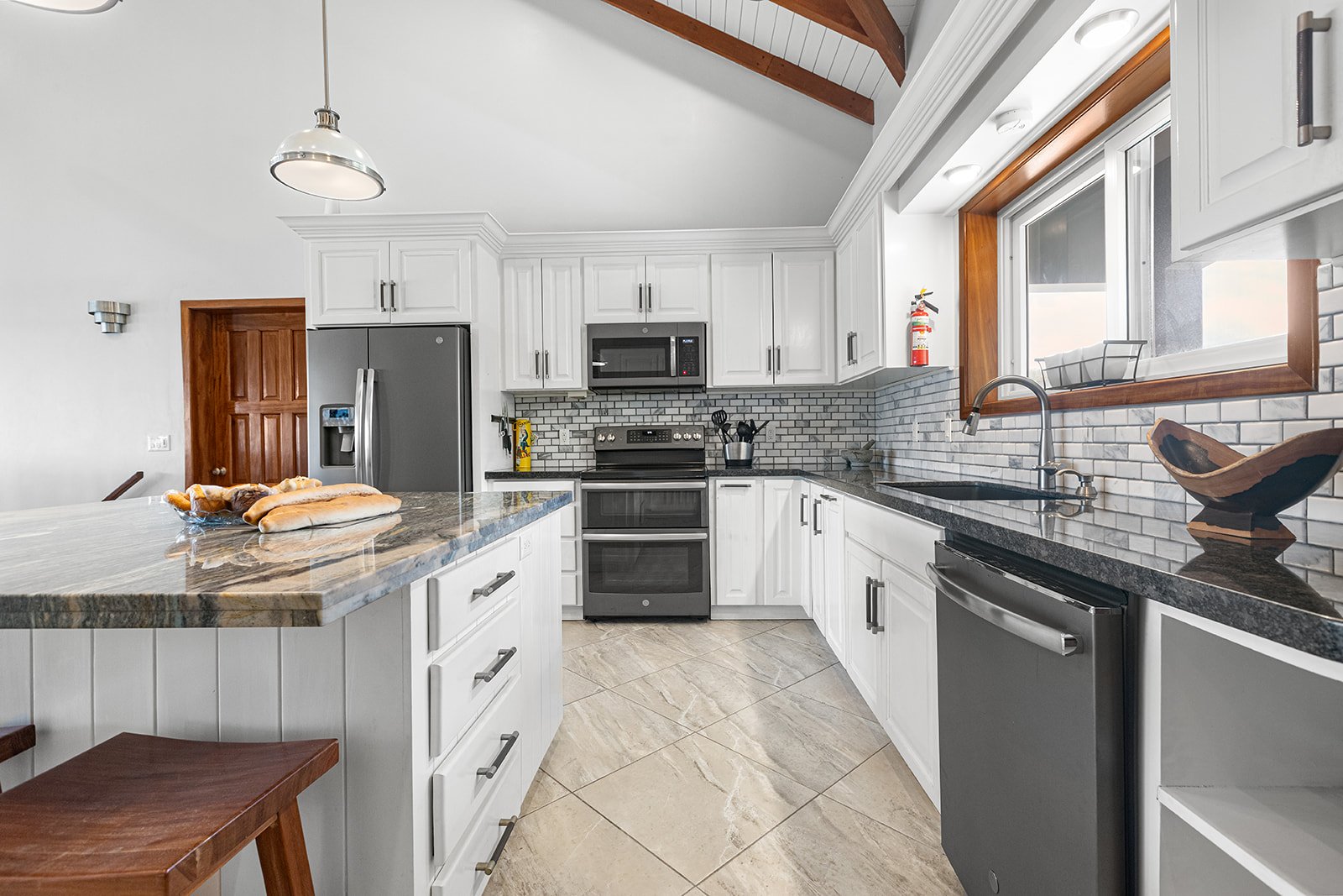 Modern kitchen with white cabinets, gray appliances, black countertops, and a marble island. The window has a wooden frame and a fire extinguisher is mounted near it.