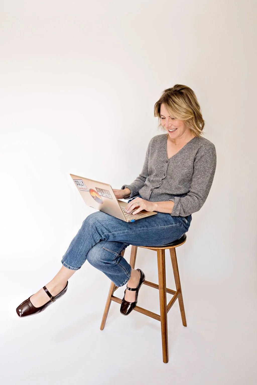 Brigette Zorn, strategic communications advisor, smiling while seated on a wooden stool and working on a laptop against a white background.