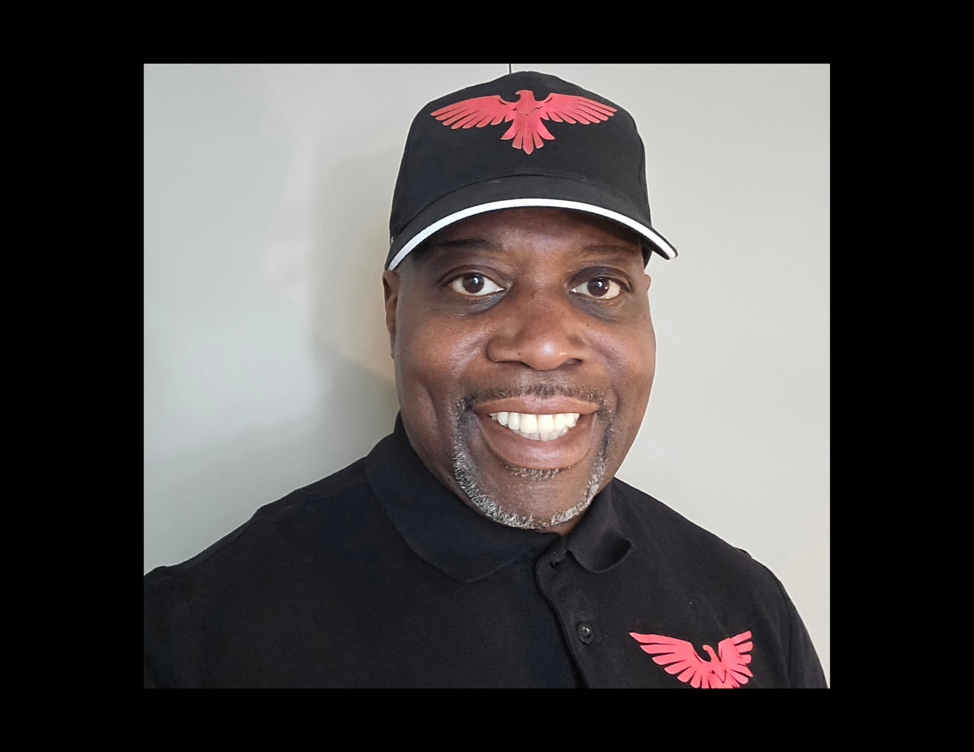 Man standing inside a partially constructed room with exposed insulation and wood framing, wearing a black shirt and cap, with a small microphone clipped to his shirt.