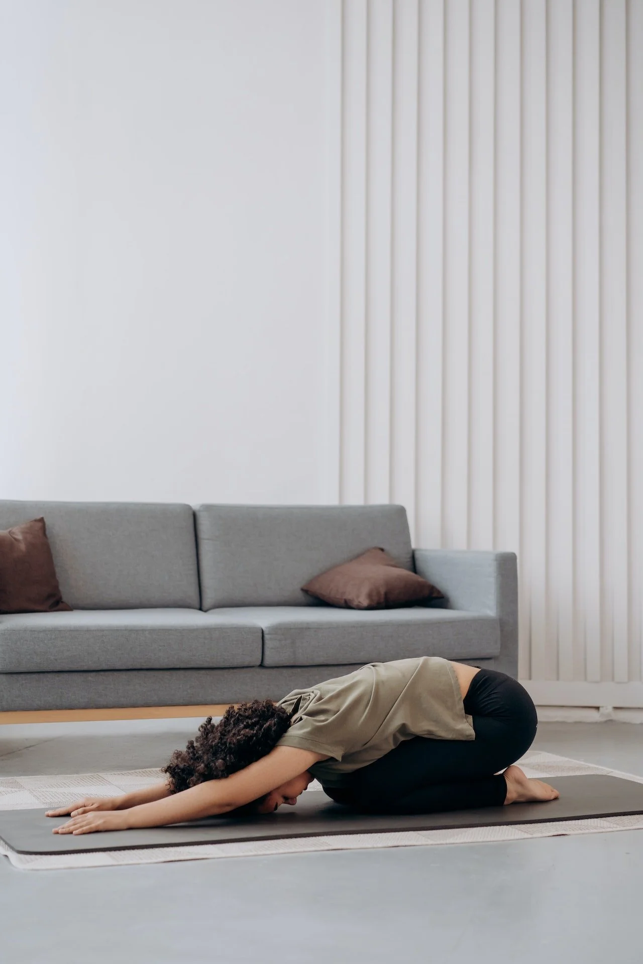 A woman practicing yoga in Child's Pose on a black mat in a living room with a gray couch and brown pillows, white wall, and vertical beige blinds.