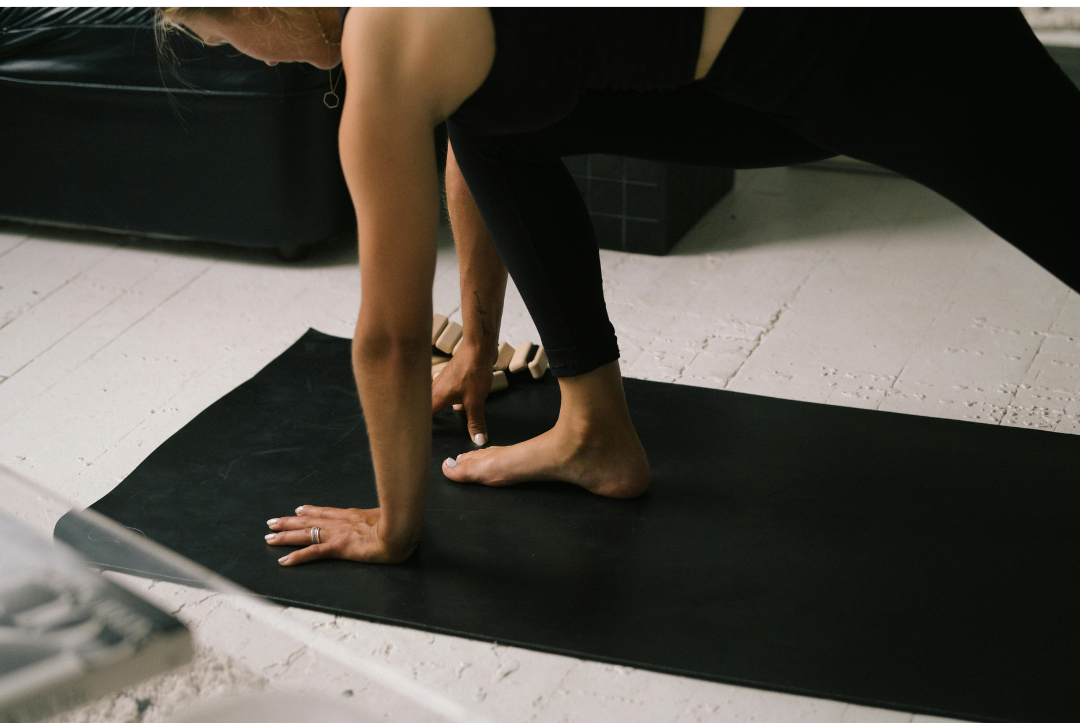 A person practicing yoga or stretching on a black yoga mat, with legs bent and hands on the mat, indoors with a white concrete floor.