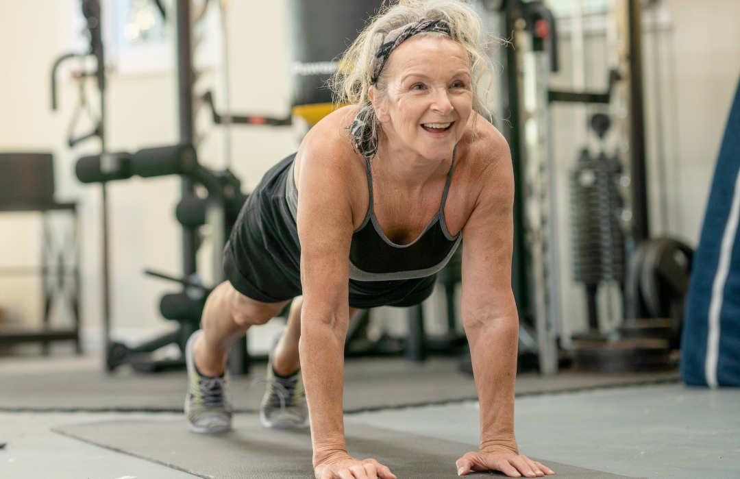 An elderly woman with gray hair in a headband doing a push-up in a gym