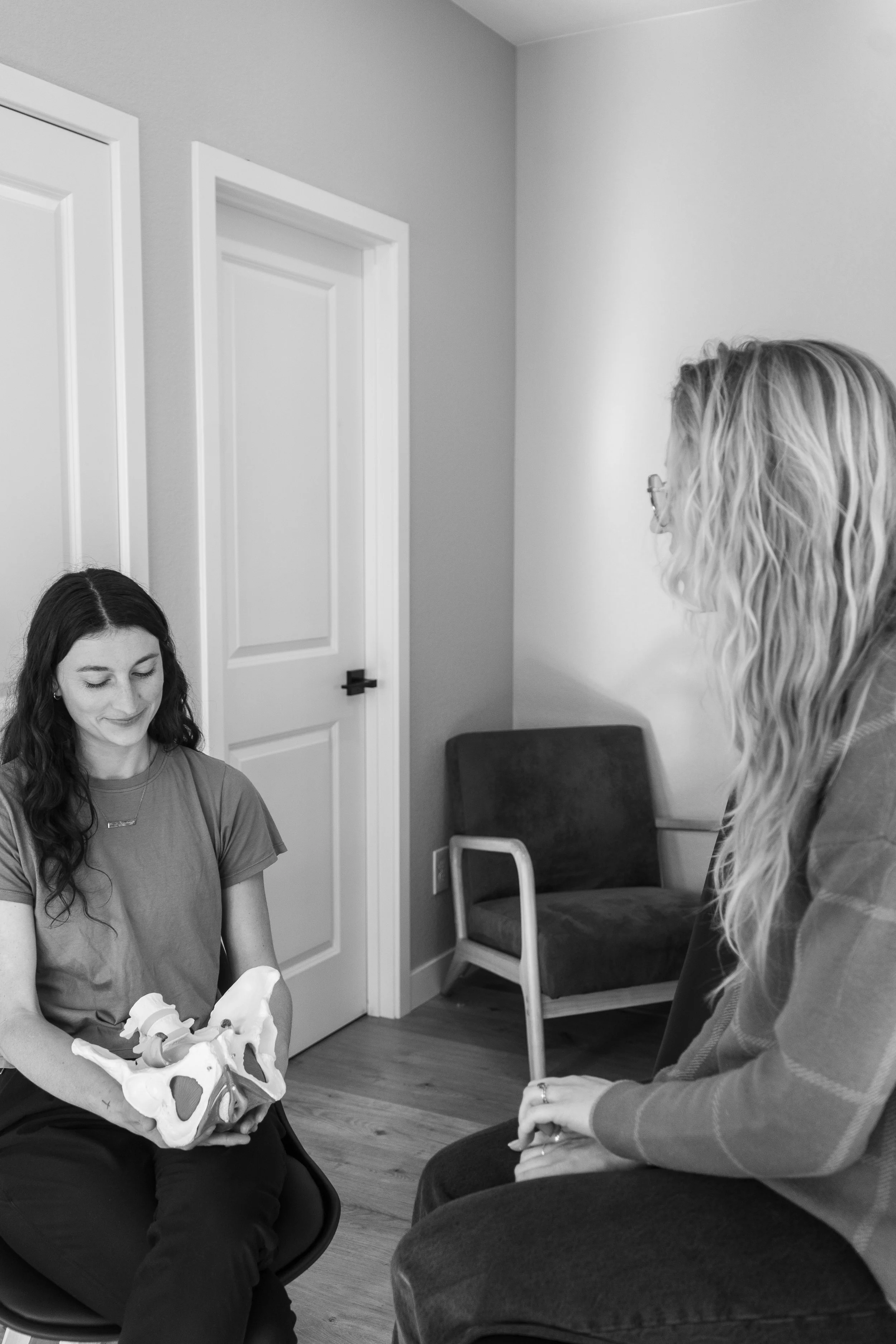 A woman sitting in a chair holding a pelvis model, talking to another woman seated facing her, in a room with a closed door and a chair in the background.