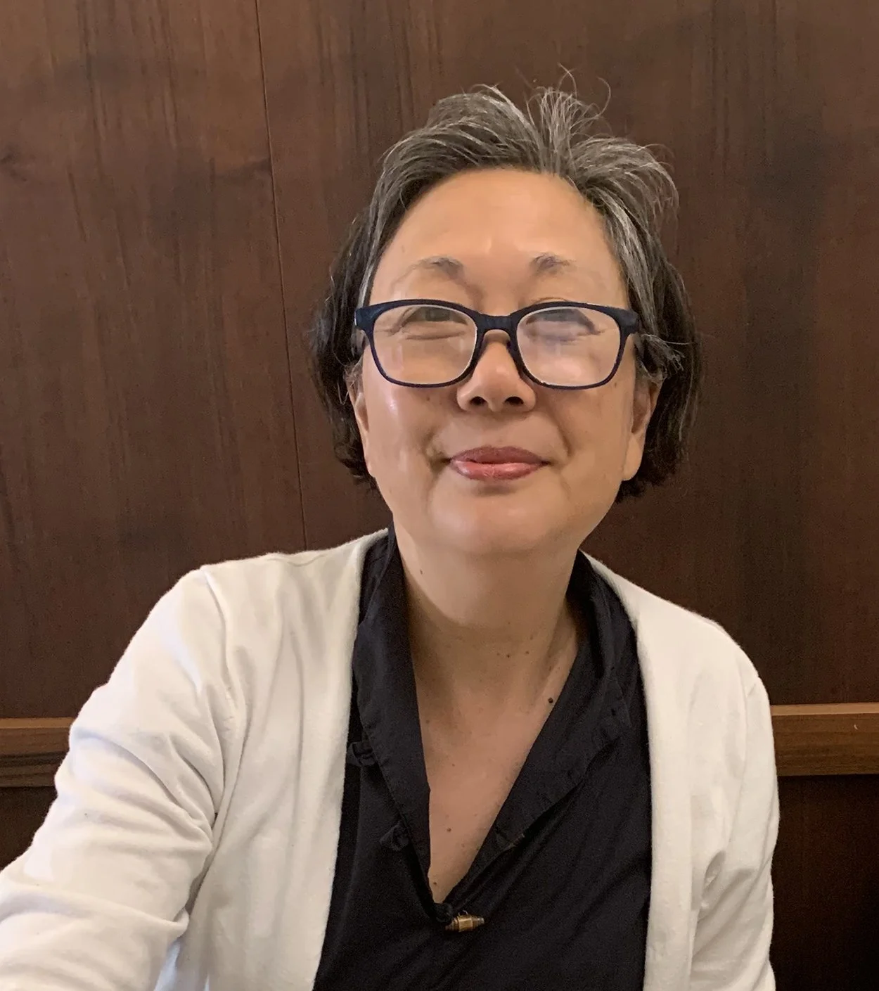 A woman with short, gray hair and glasses making a playful face with her tongue slightly out. She is wearing a white cardigan over a black top and is sitting against a wooden background.