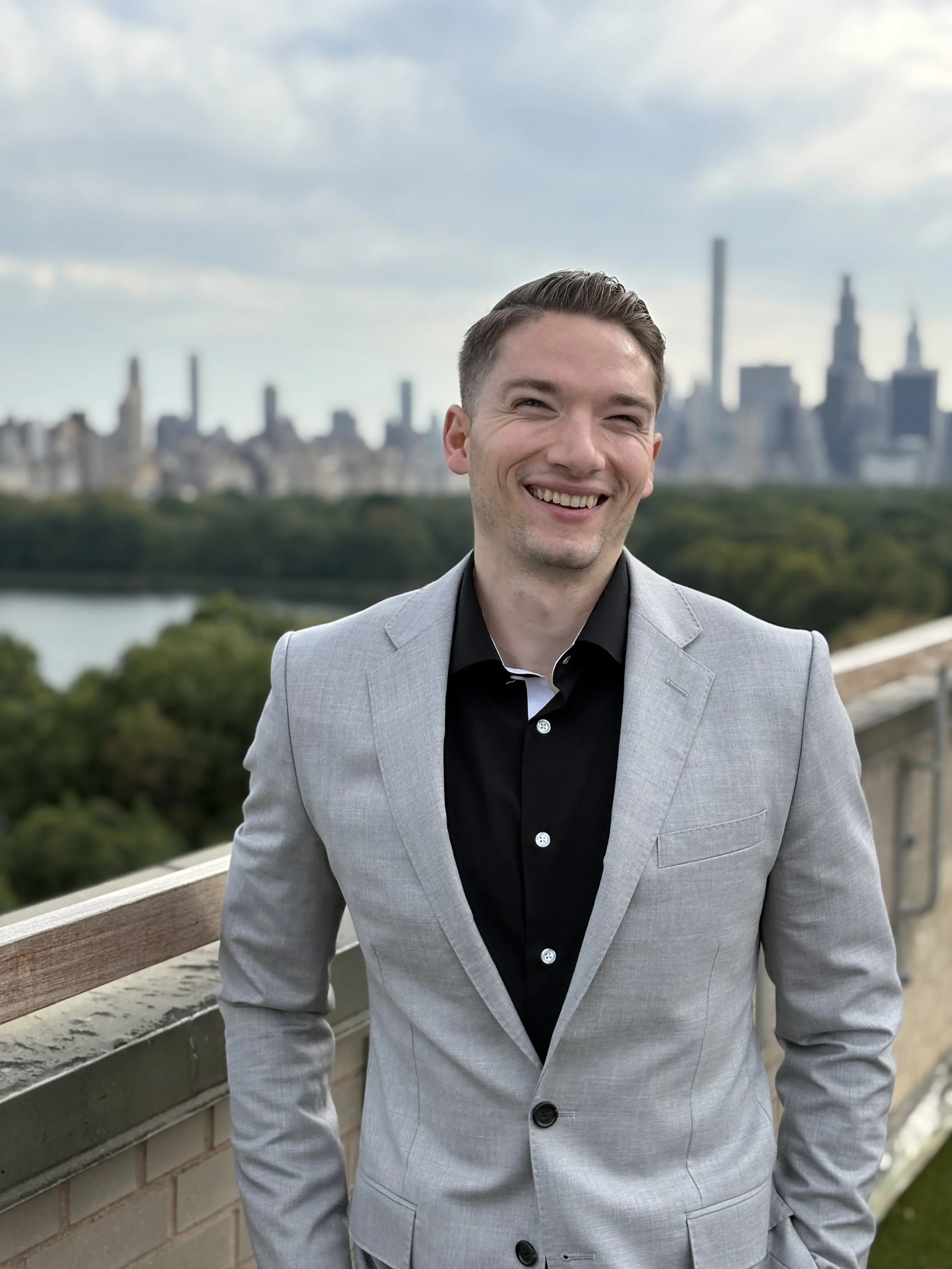 A smiling man in a light gray suit with a black shirt standing outdoors on a balcony, with a city skyline, trees, and a river in the background.