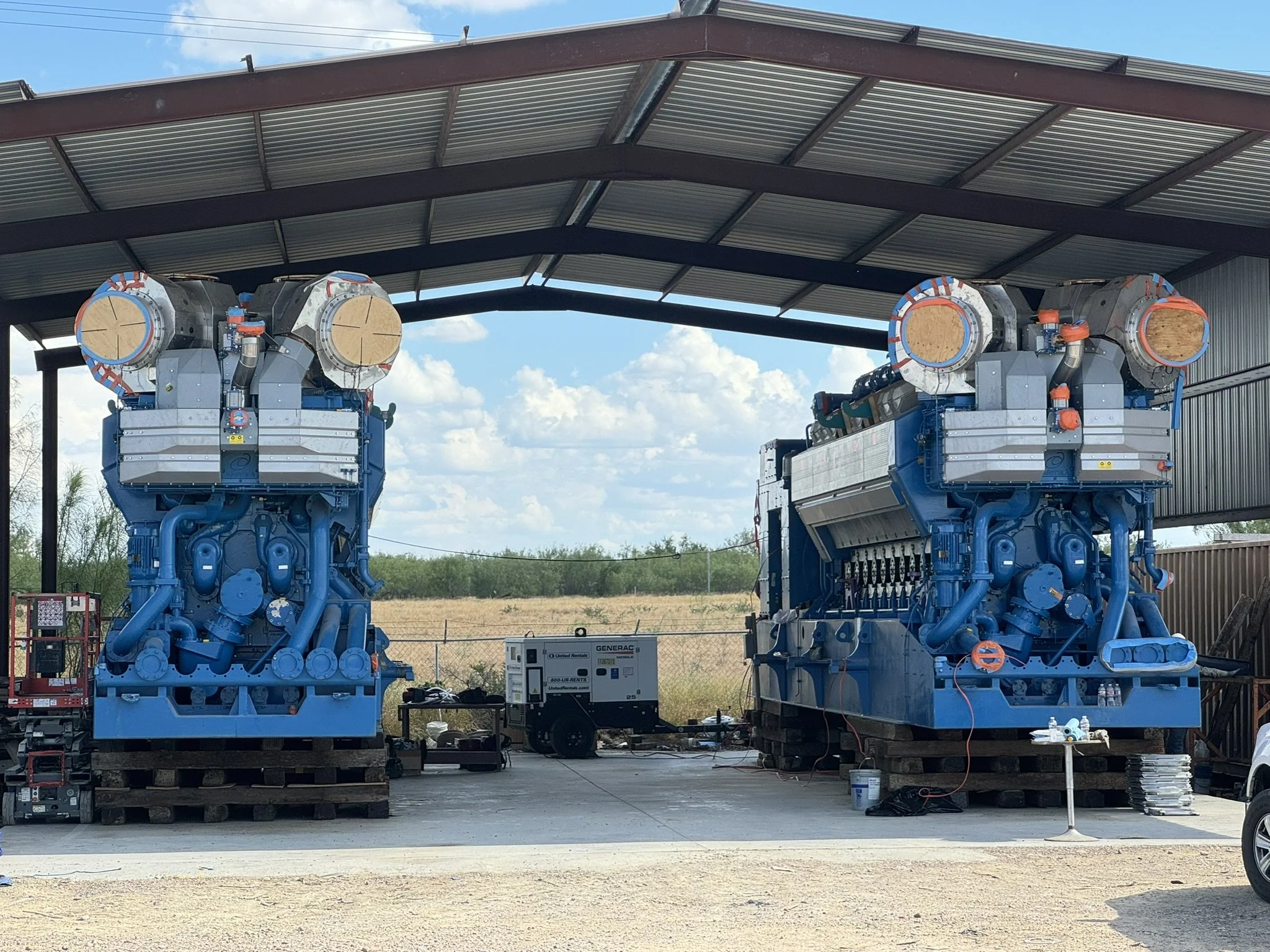 Two large blue industrial engines or turbines under a metal shelter, with desert landscape and blue sky in the background.