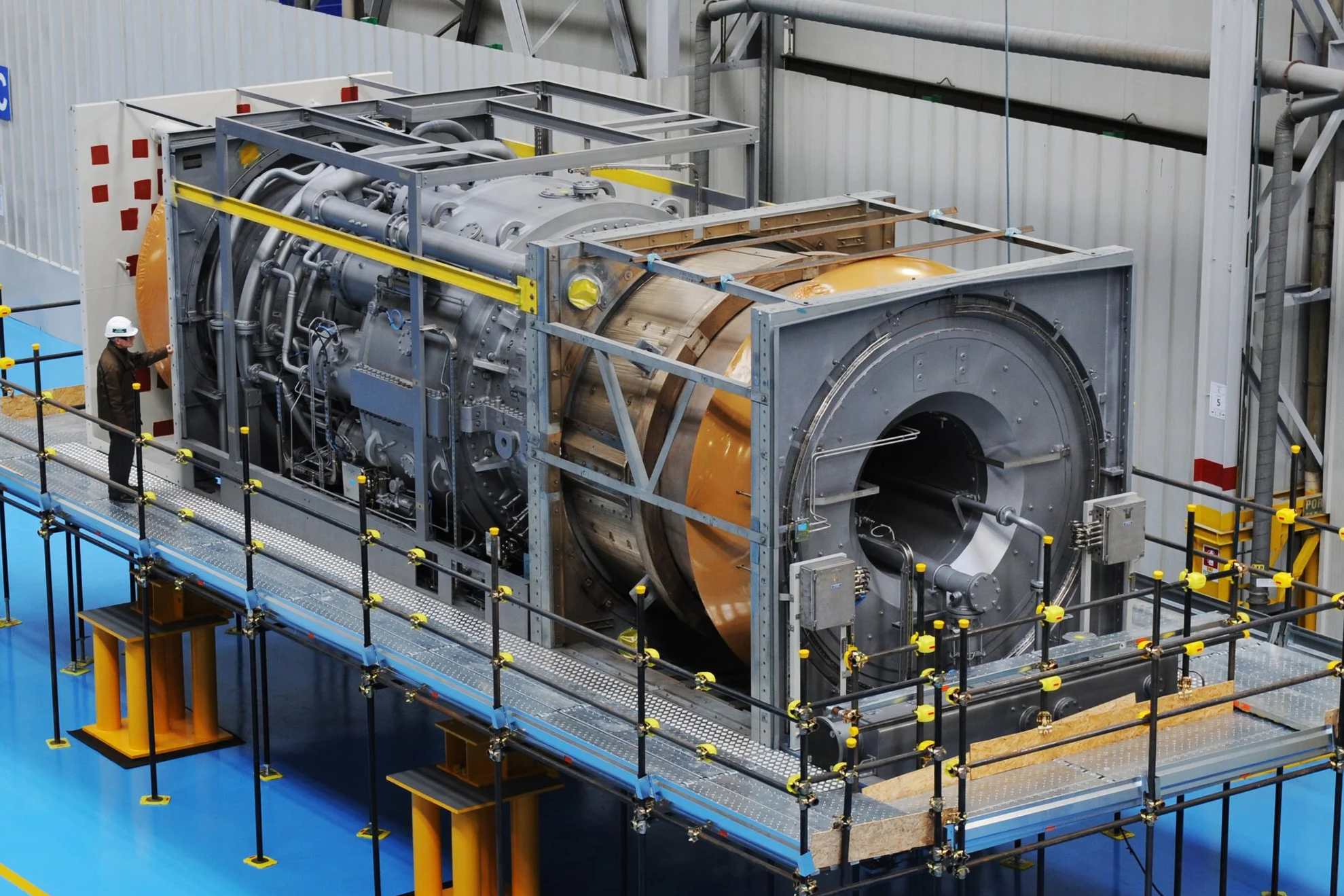 Large industrial machine with various pipes and components, surrounded by safety barriers, in a factory setting, with a worker observing.