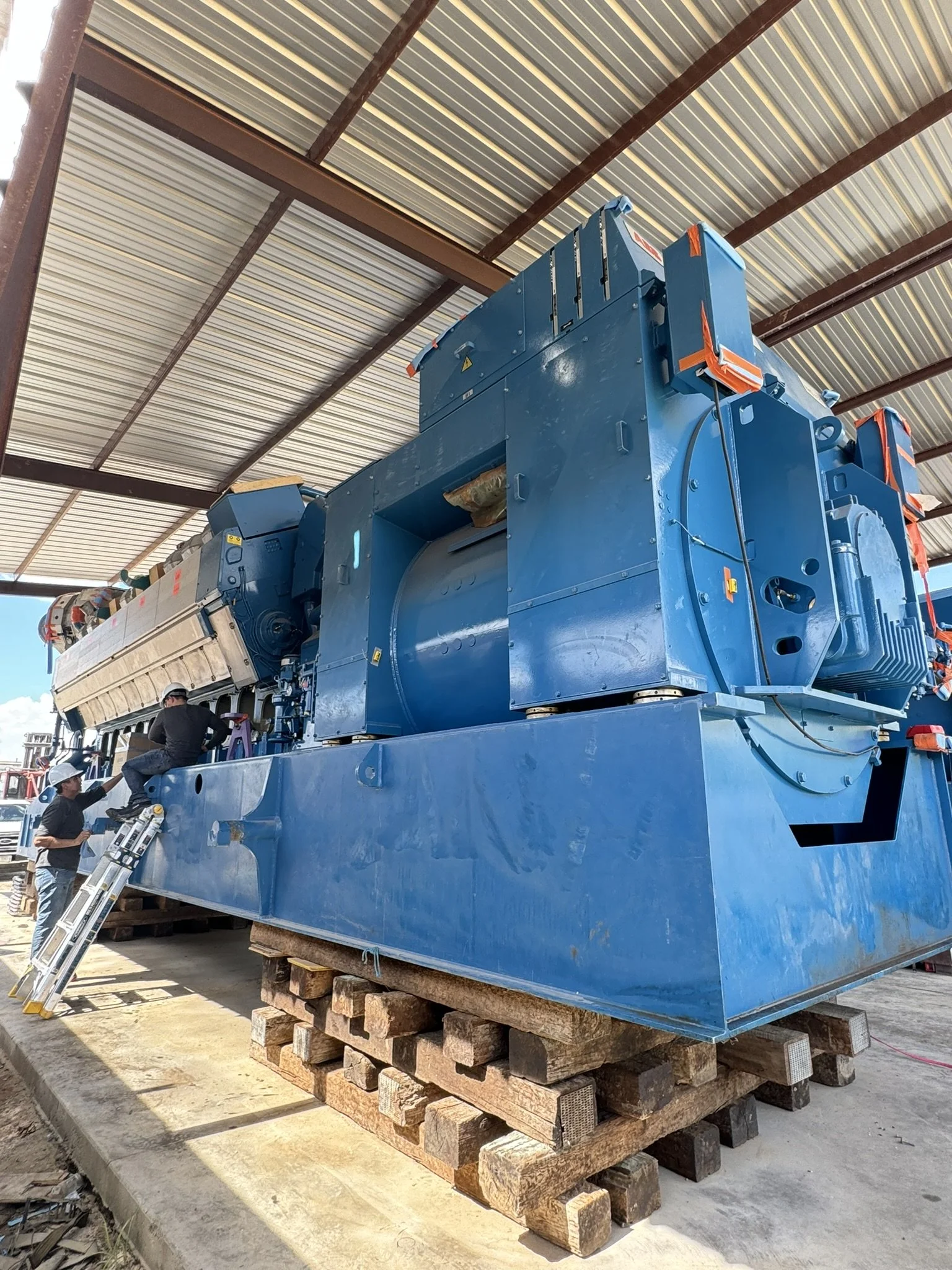 Large industrial blue machine in a warehouse, with workers inspecting it, placed on wooden pallets.