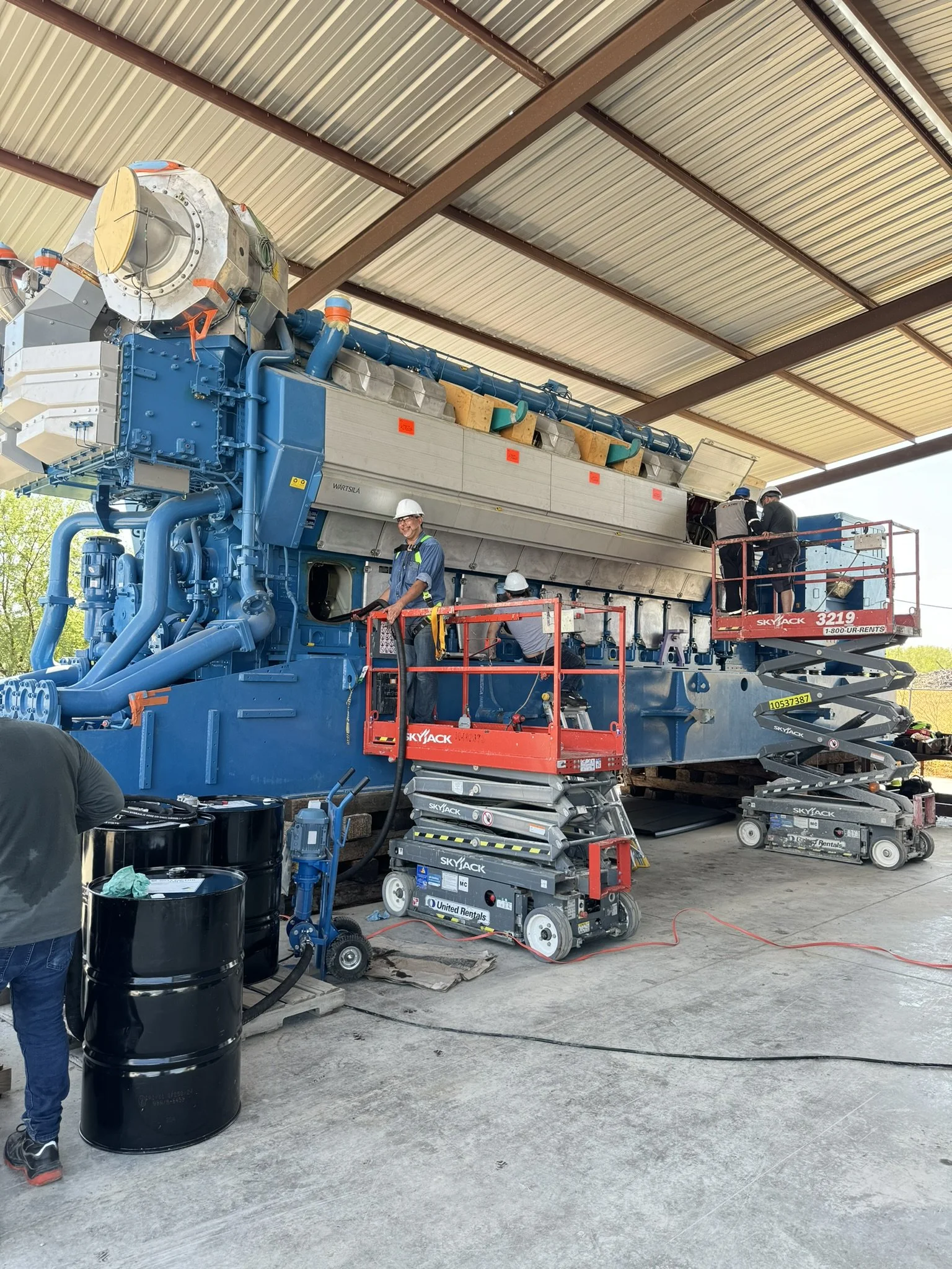 Workers inspecting a large industrial machine inside a covered area with scissor lifts and tools.