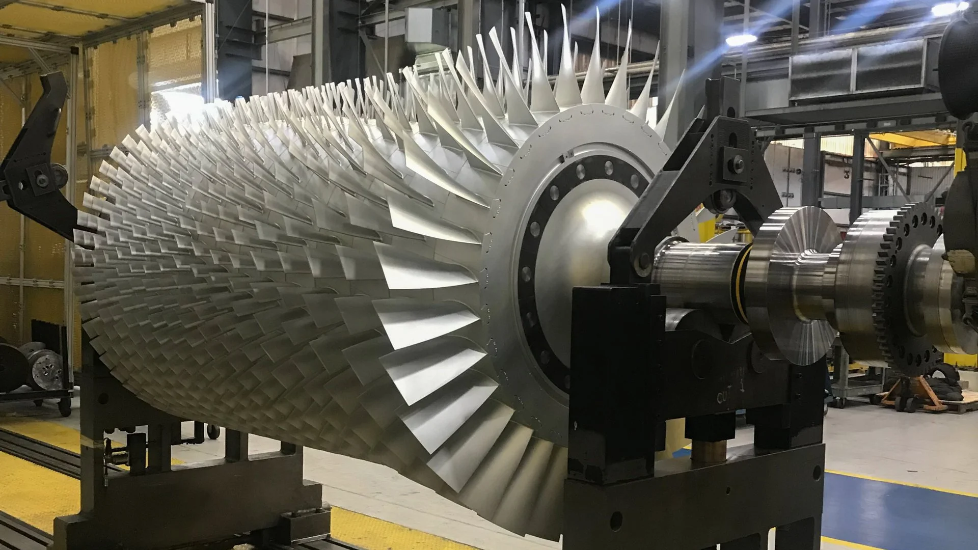 Large industrial jet engine component with numerous titanium blades mounted on a metal frame inside a manufacturing facility.