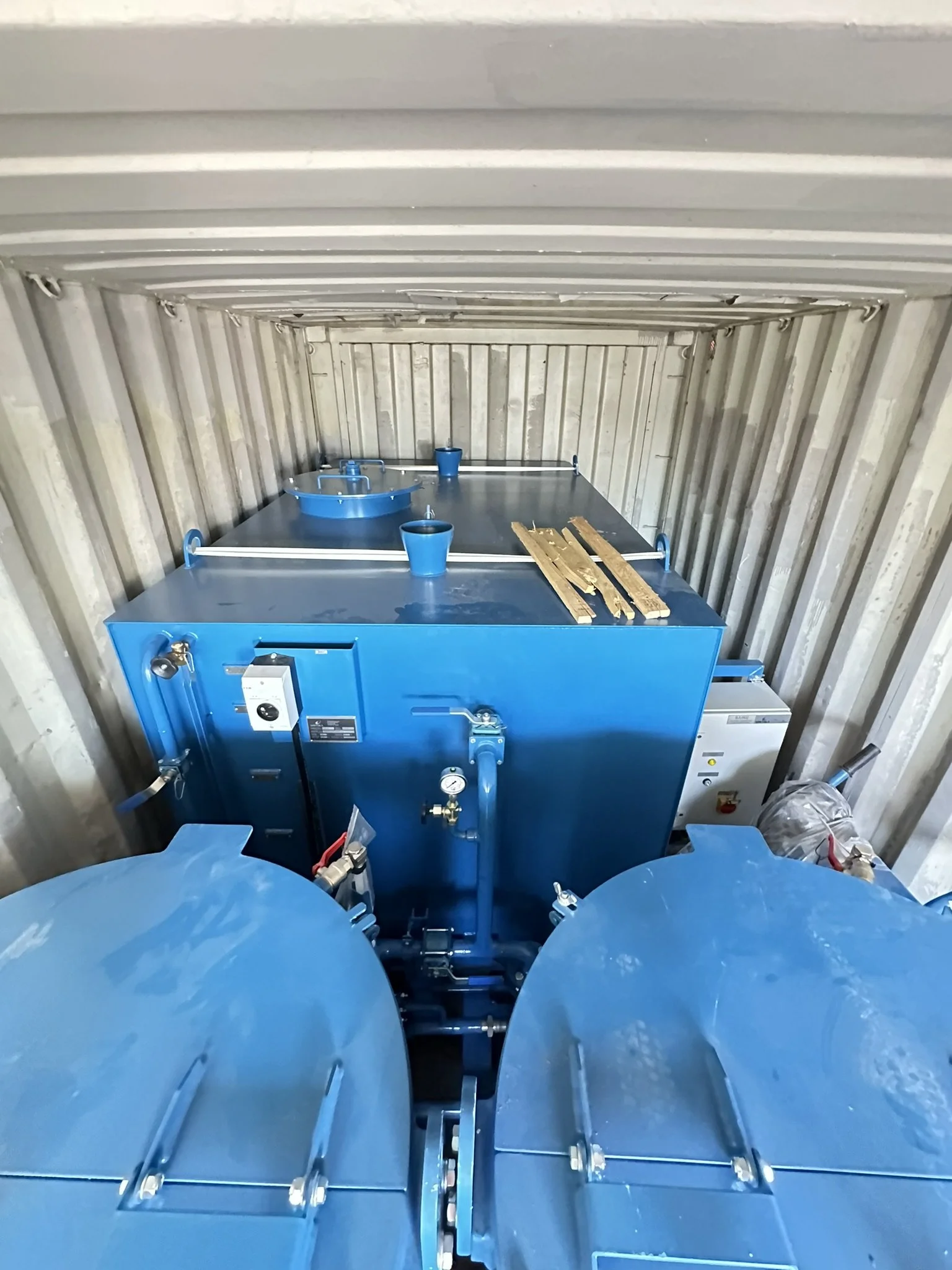 View inside a shipping container with a large blue industrial machine, two blue cups, two wooden pallets, and a small white box on the right side.