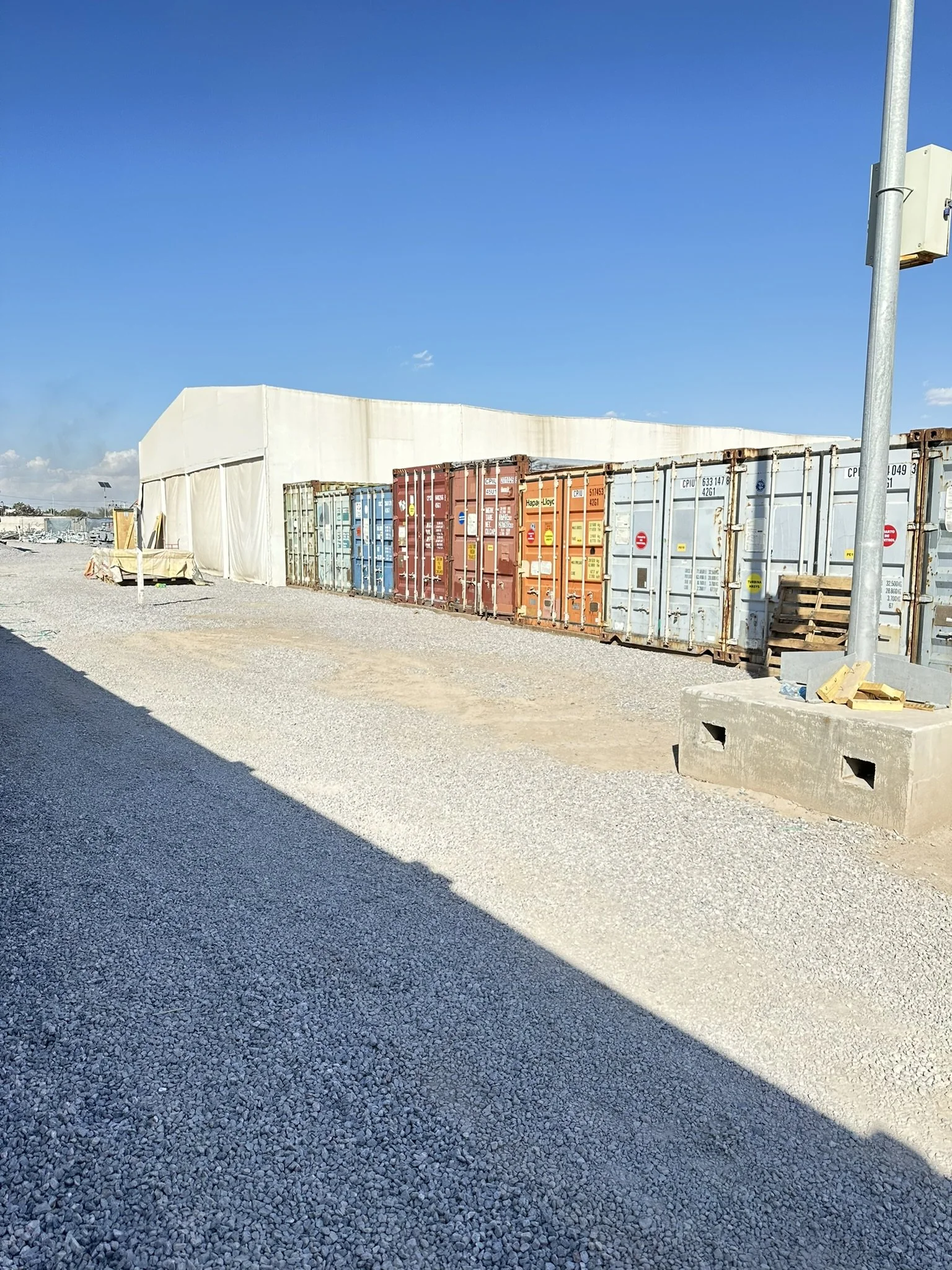Row of colorful shipping containers beside a large white tent structure on a gravel lot under a clear blue sky.