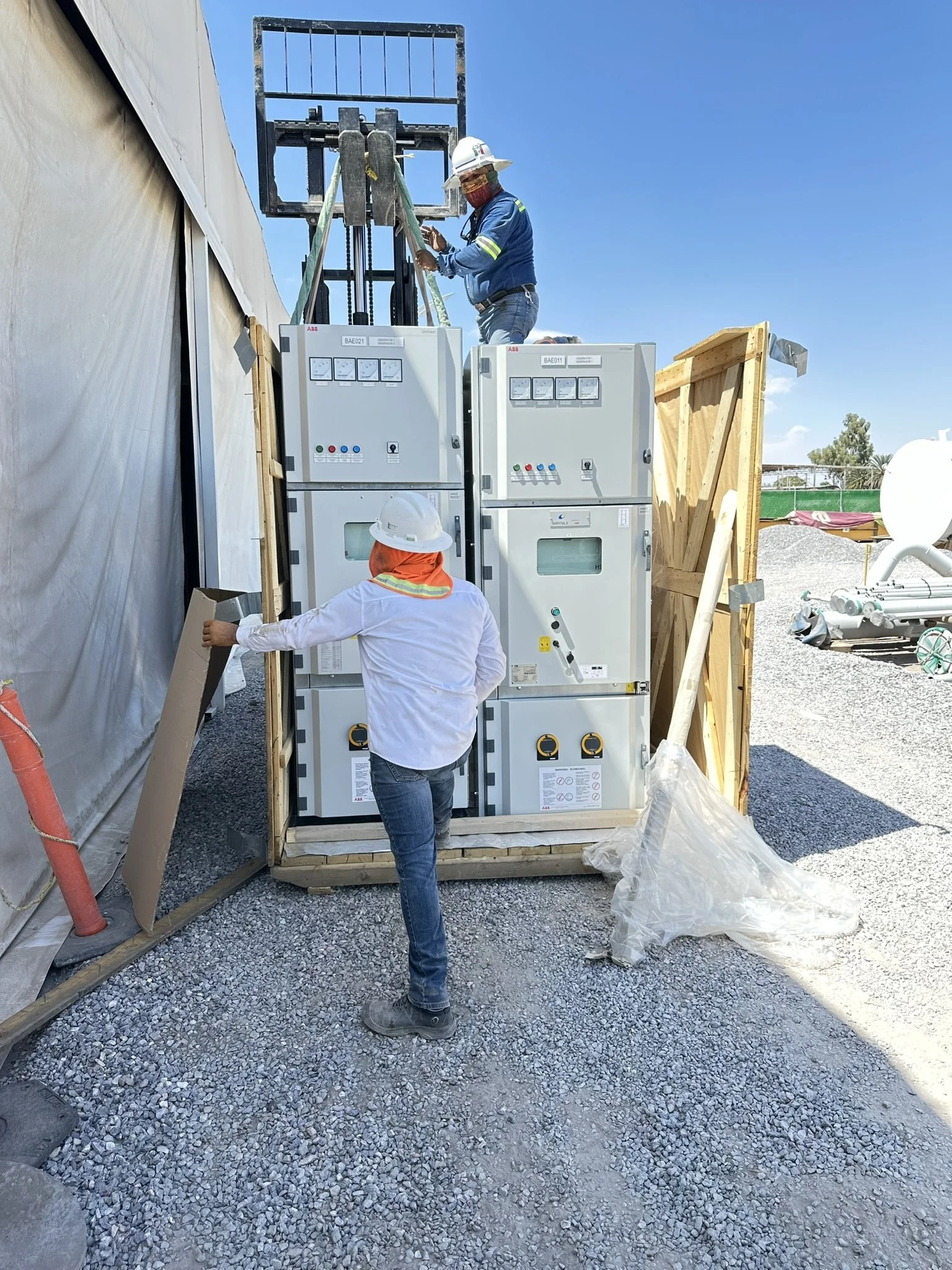 Three workers wearing safety gear and hard hats are working with electrical equipment outdoors during the daytime. One worker is standing on top of a large electrical panel, while another is standing nearby holding a cardboard piece. The third worker is observing and possibly assisting.