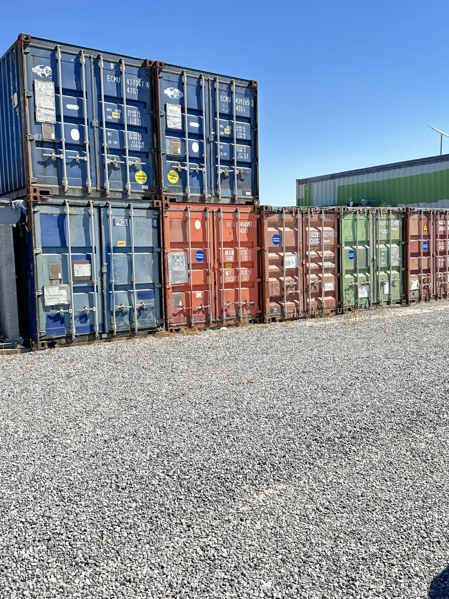 Several colorful shipping containers stacked on a gravel lot against a clear blue sky.