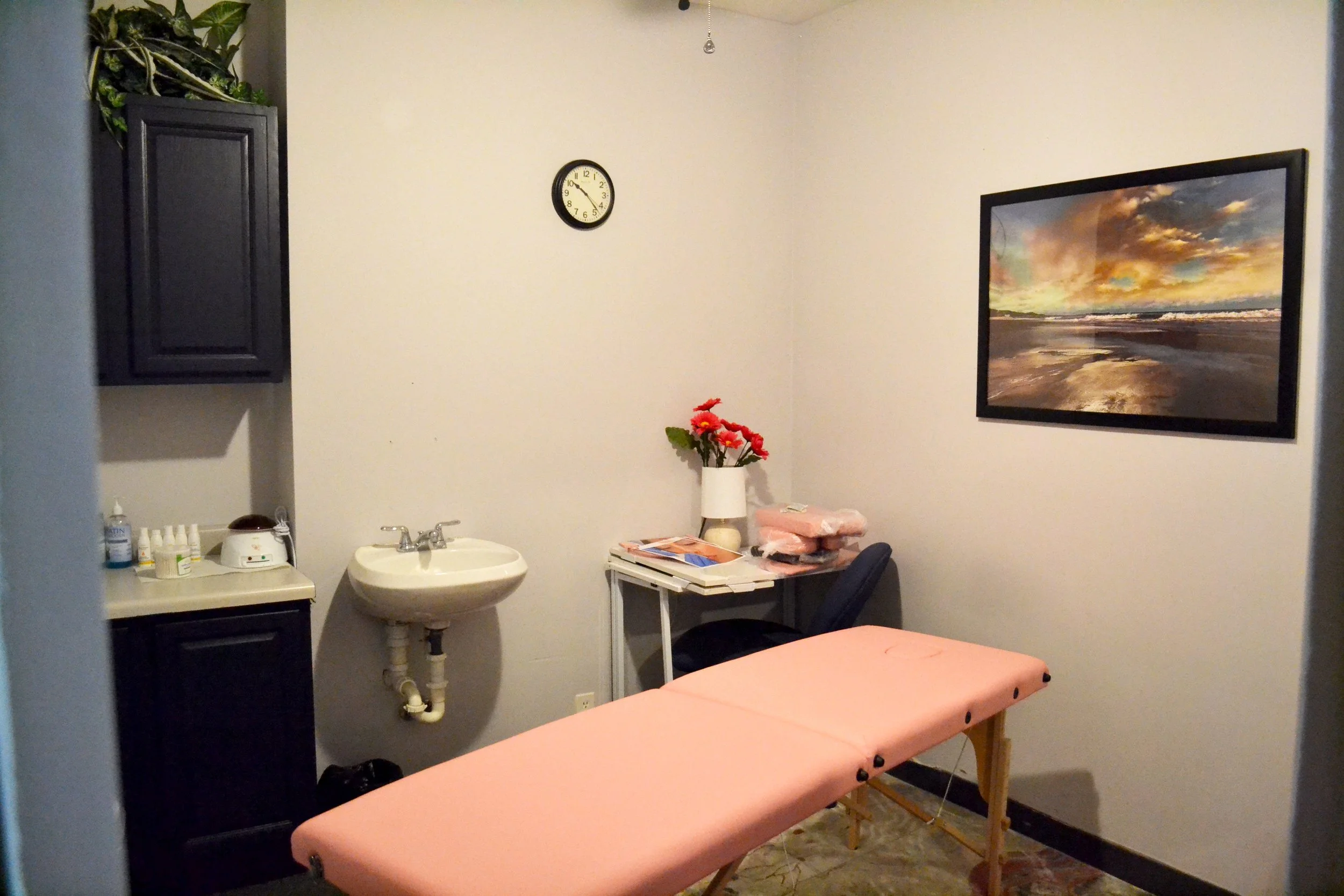 Massage therapy room with a pink massage table, a white sink, a black cabinet, a desk with a vase of red flowers, a chair, and framed landscape photo on the wall.