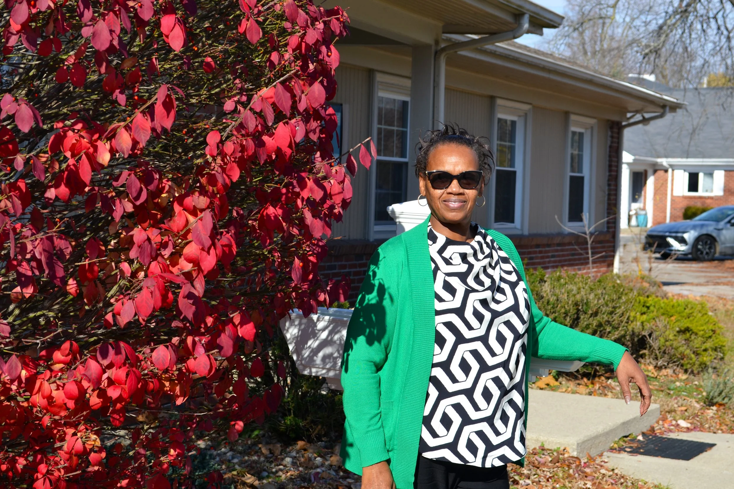 A woman with sunglasses standing outside in front of a house with red and pink leaves on a bush.