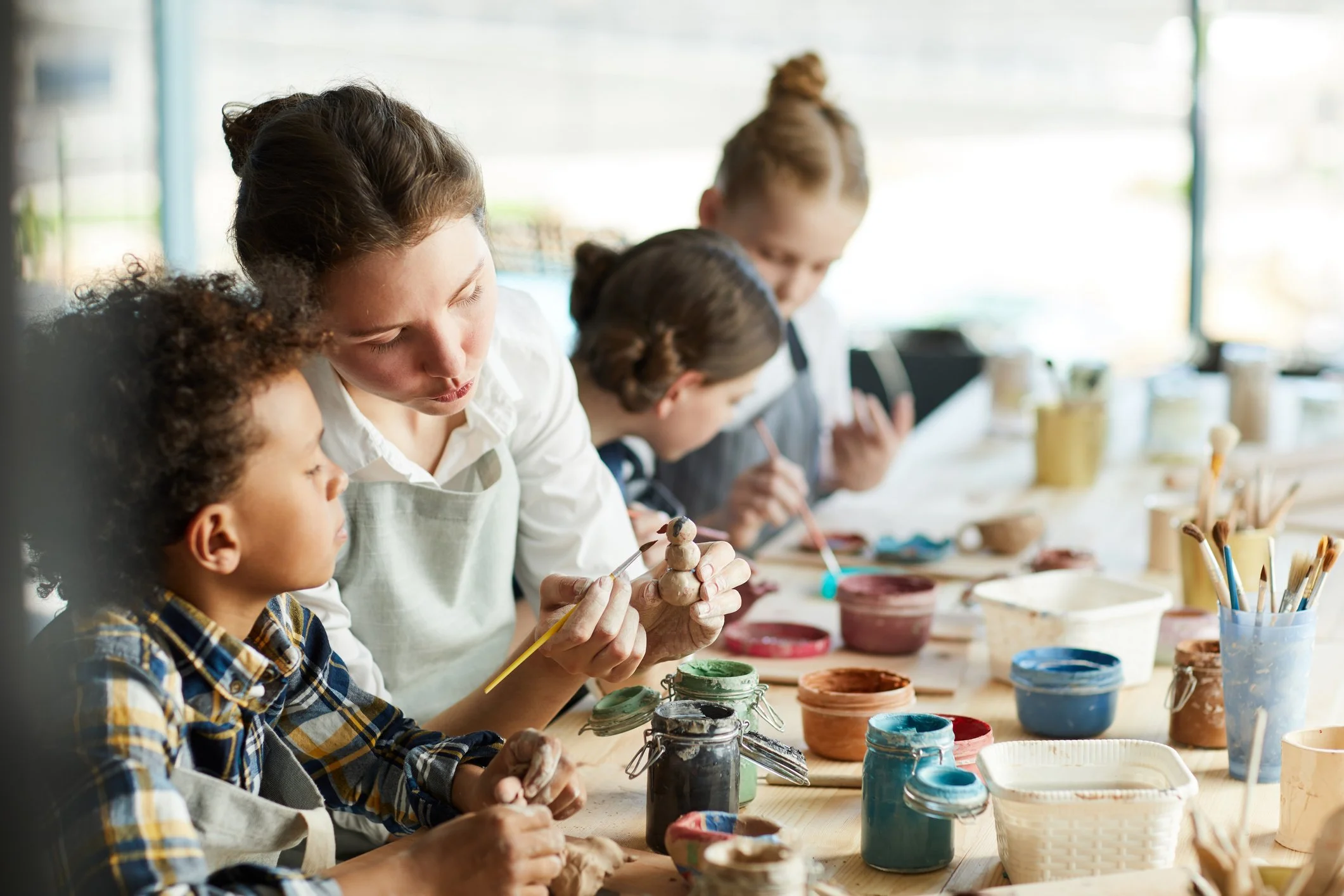 Children and an instructor painting ceramics in a studio with art supplies on a wooden table.