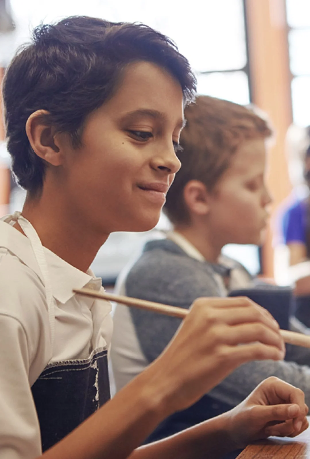 A boy with dark hair and light skin holds chopsticks and smiles while sitting at a table in a classroom or dining area, with other children in the background.
