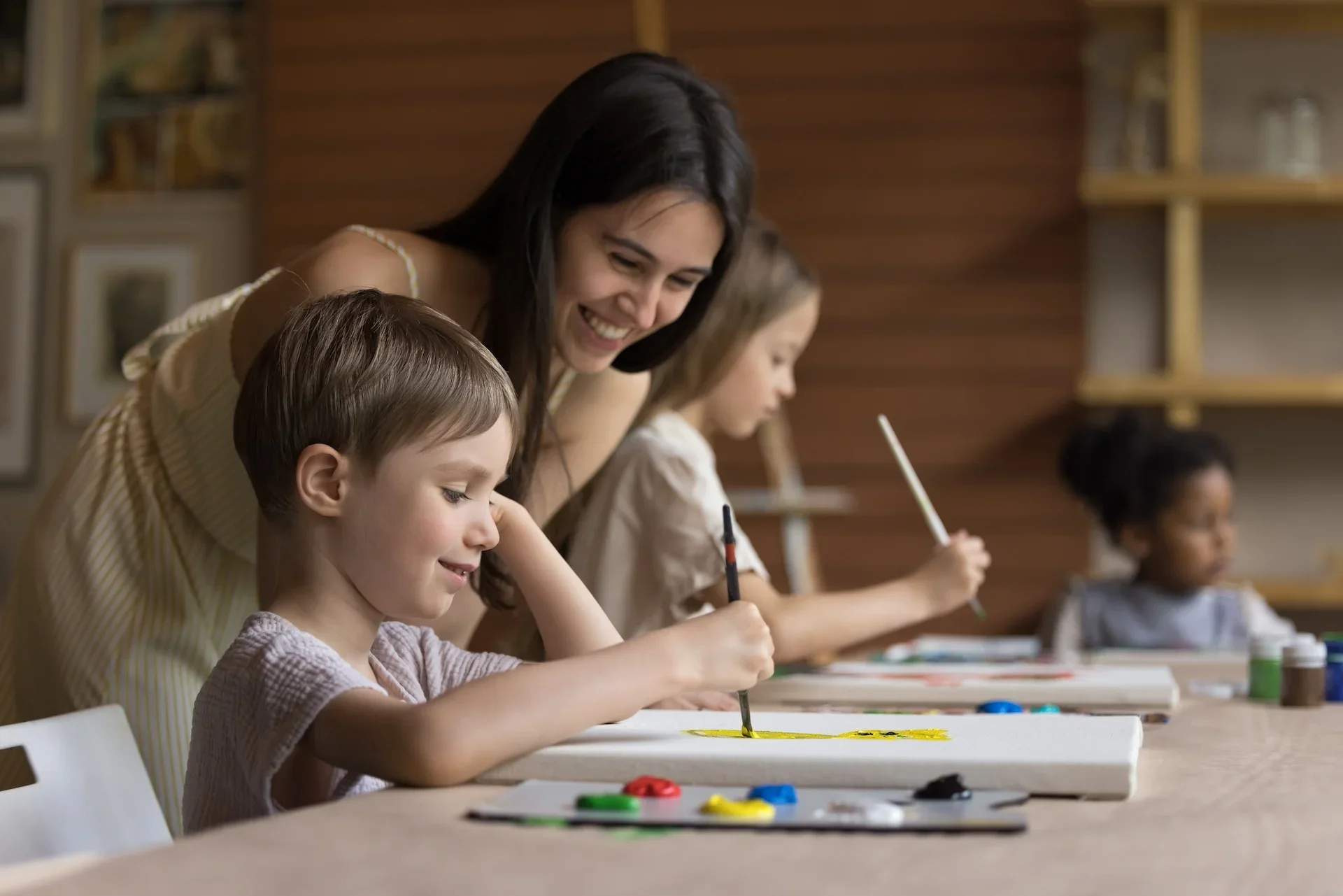 A woman and three children participating in a painting activity at a table, with paintings and paint supplies in front of them.