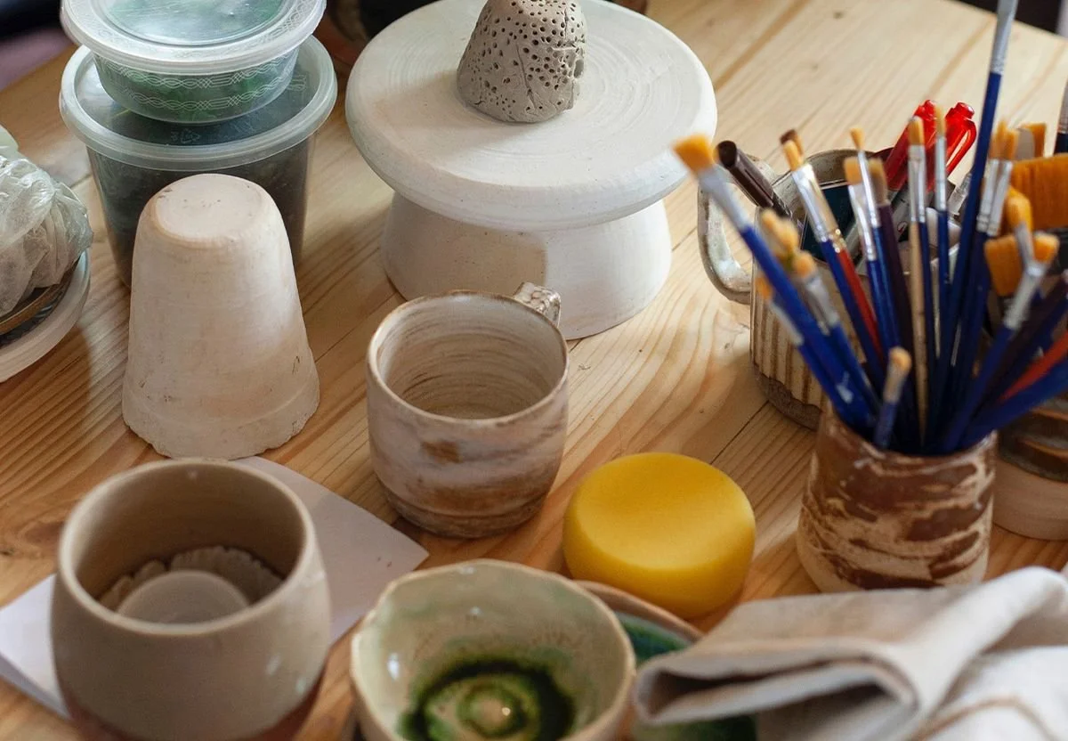 Collection of pottery tools and supplies including cups, bowls, a sponge, and paintbrushes on a wooden table.