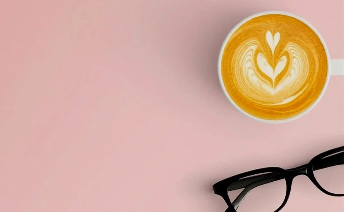 A cup of coffee with latte art in the shape of a heart and a pair of black eyeglasses on a light pink surface.