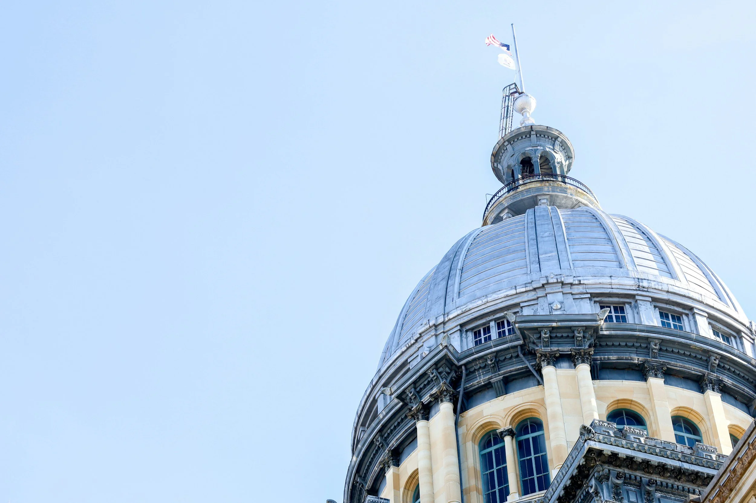 Close-up of a historic dome with a flagpole and flags on top, on a bright day with blue sky.