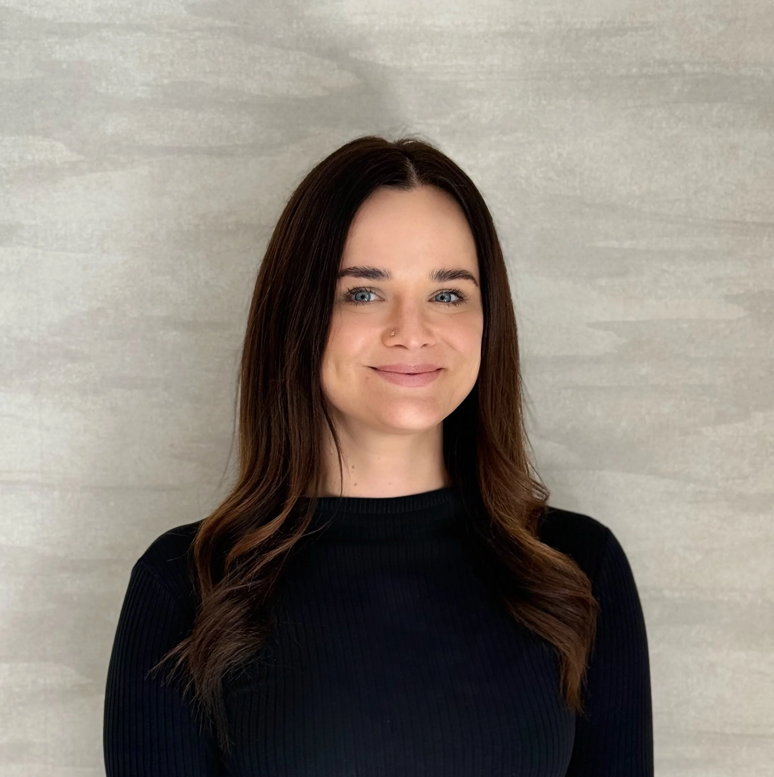 A woman with long dark brown hair, blue eyes, and a nose piercing, smiling, wearing a black top, standing against a light gray background.