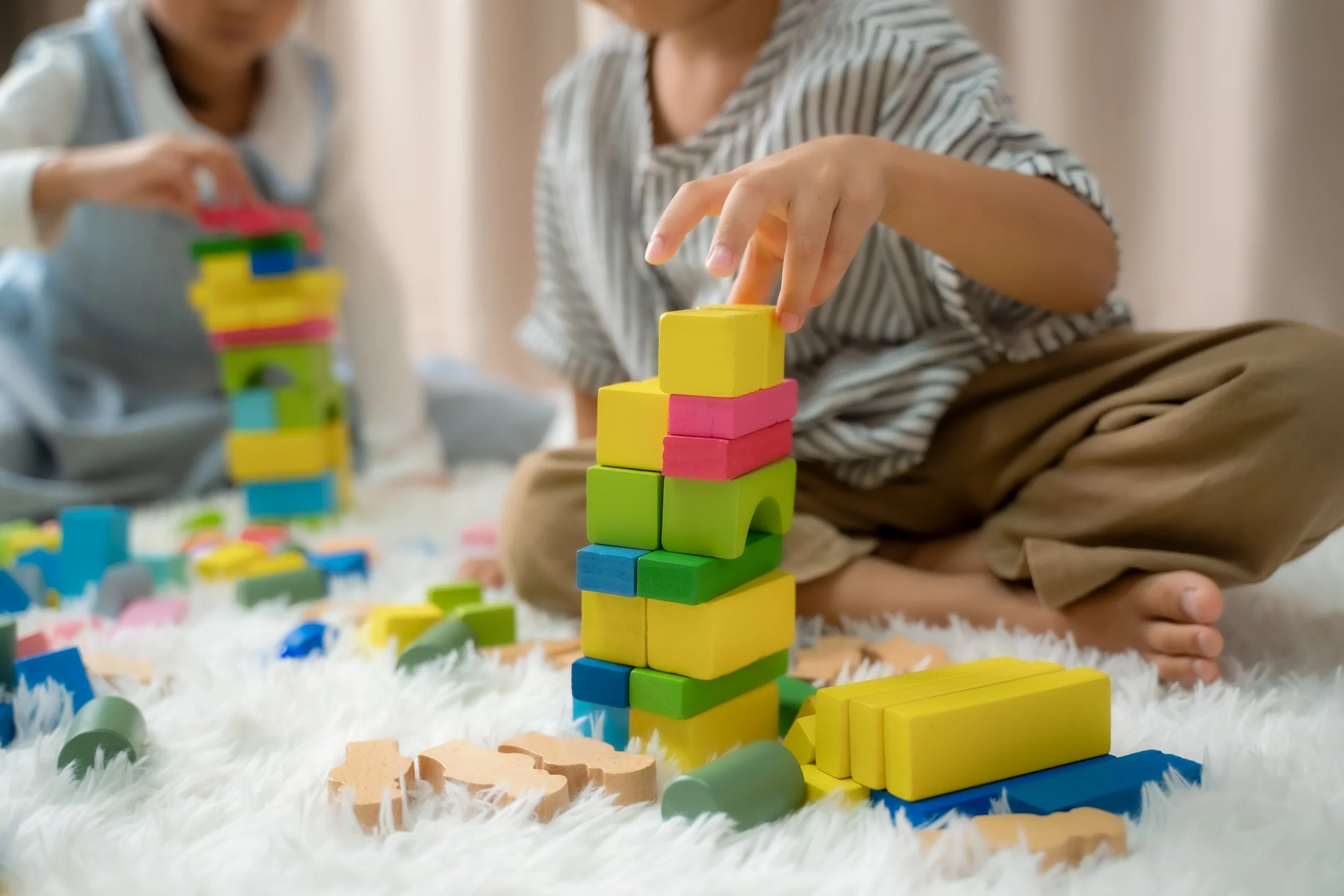 Children playing with colorful wooden building blocks on a white fluffy rug.