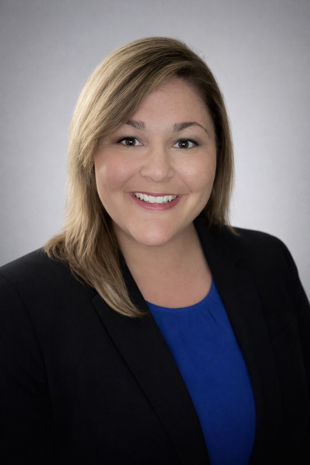 Headshot of a woman with shoulder-length blonde hair, smiling, wearing a black blazer and blue top against a plain gray background.