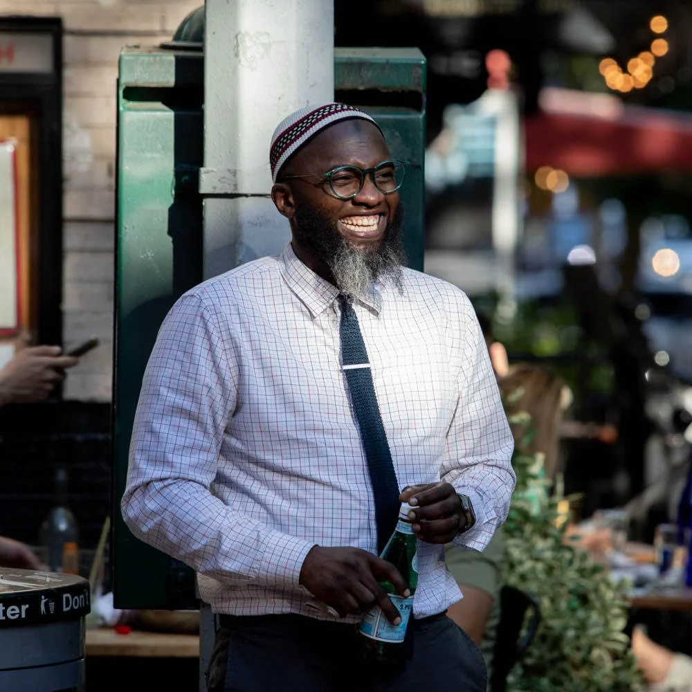 A man smiling and holding a drink outside, wearing glasses, a checkered shirt, and a black tie, with a beard and a traditional cap, in a lively outdoor setting with blurred lights and people in the background.