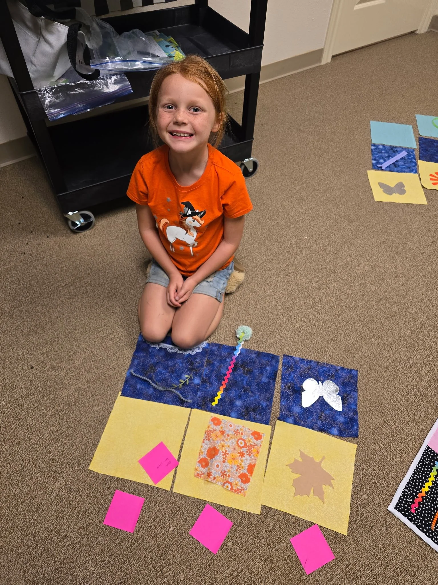 A girl with red hair, wearing an orange T-shirt with a witch hat and a white horse on it, is kneeling on the carpet in a room. In front of her, there are six colored papers arranged on the floor, with three pink sticky notes placed at the bottom. On 