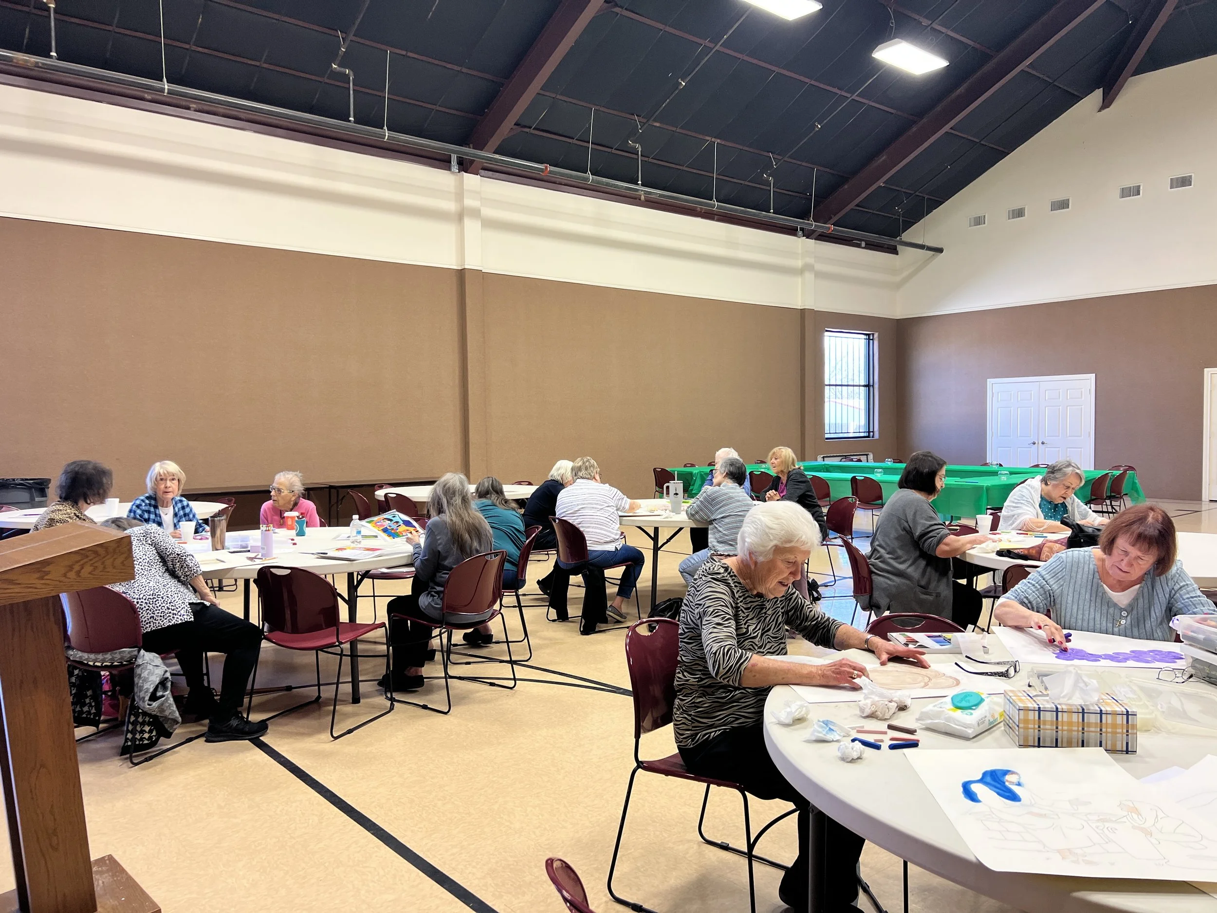 Group of elderly women engaged in arts and crafts activities at round tables inside a large room or community center.