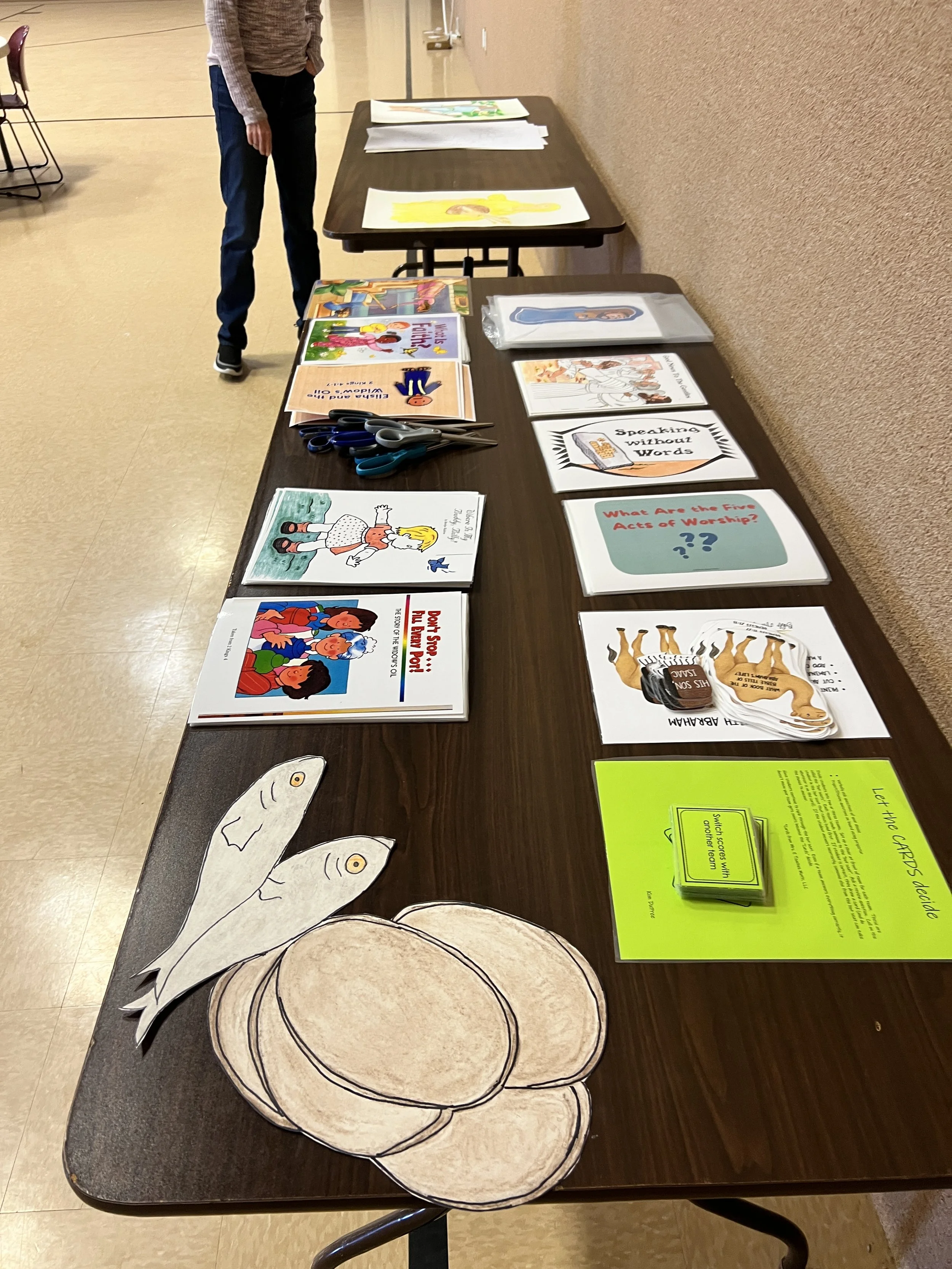 Table with children’s educational books, paper fish and golden potatoes cut-outs, scissors, and a sign about a kids' activity station.