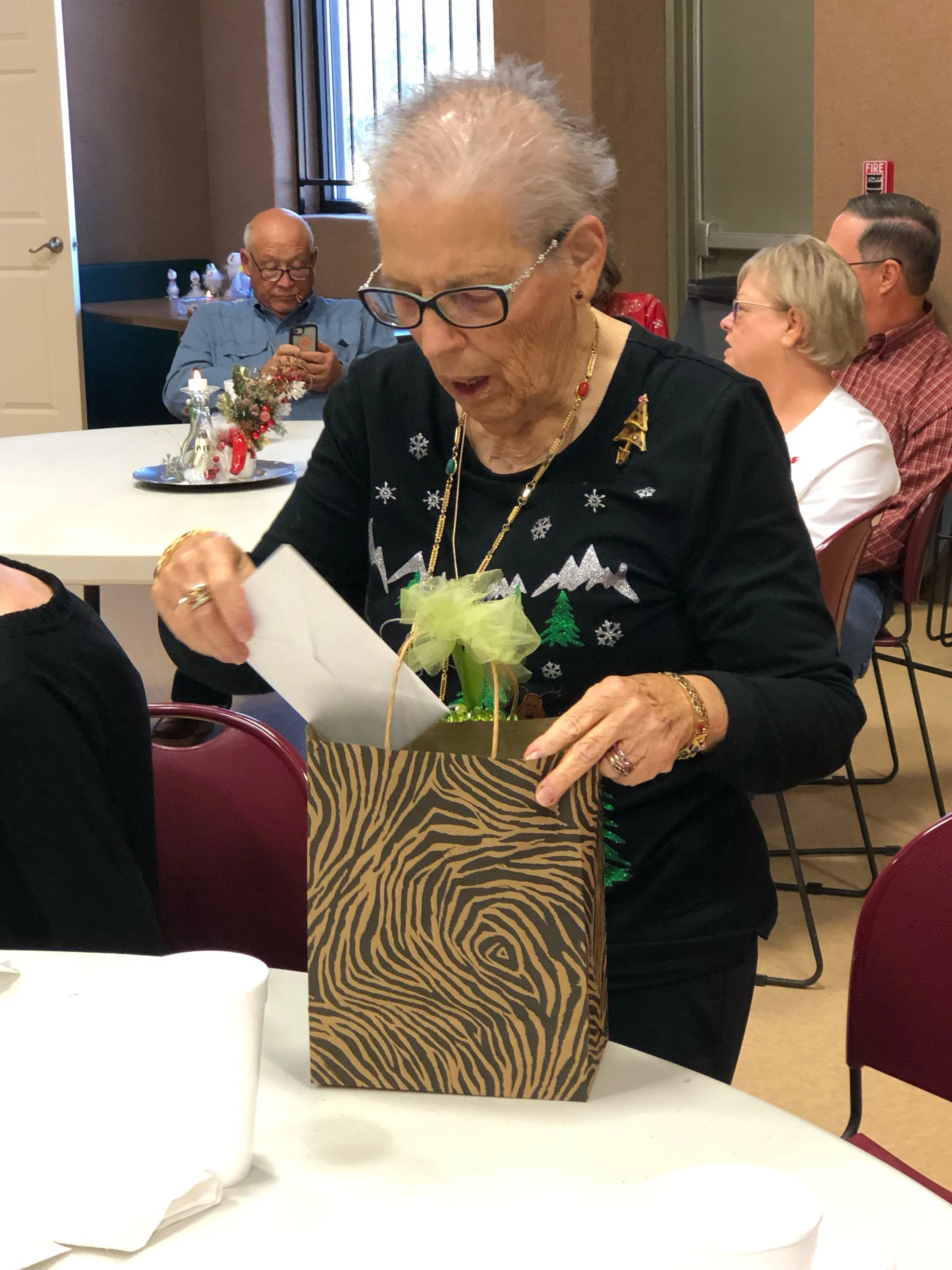 An elderly woman with glasses and holiday jewelry opening a gift bag with a surprised expression at a holiday gathering.